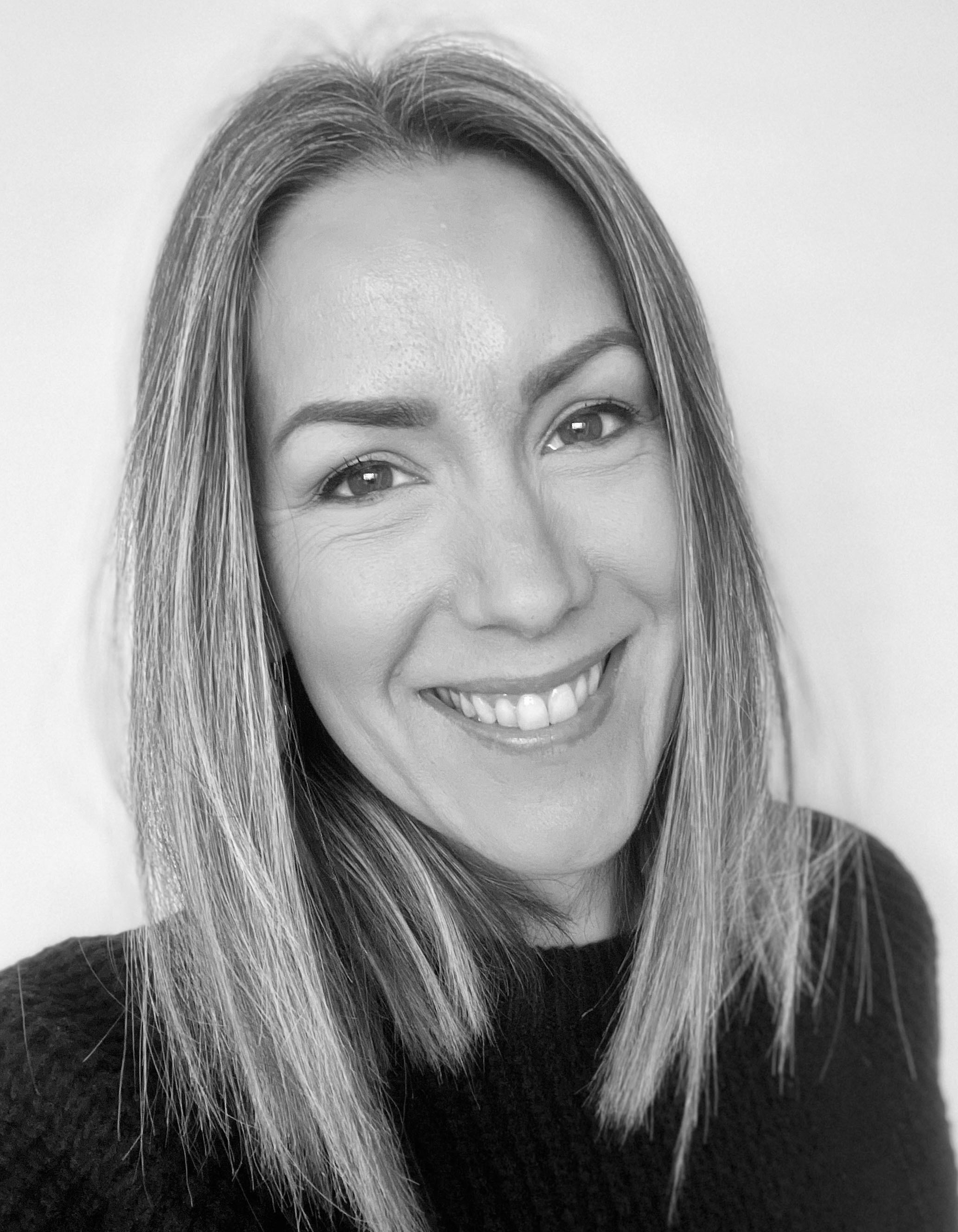 Black and white headshot of smiling woman with shoulder-length blonde hair wearing dark top against light background.