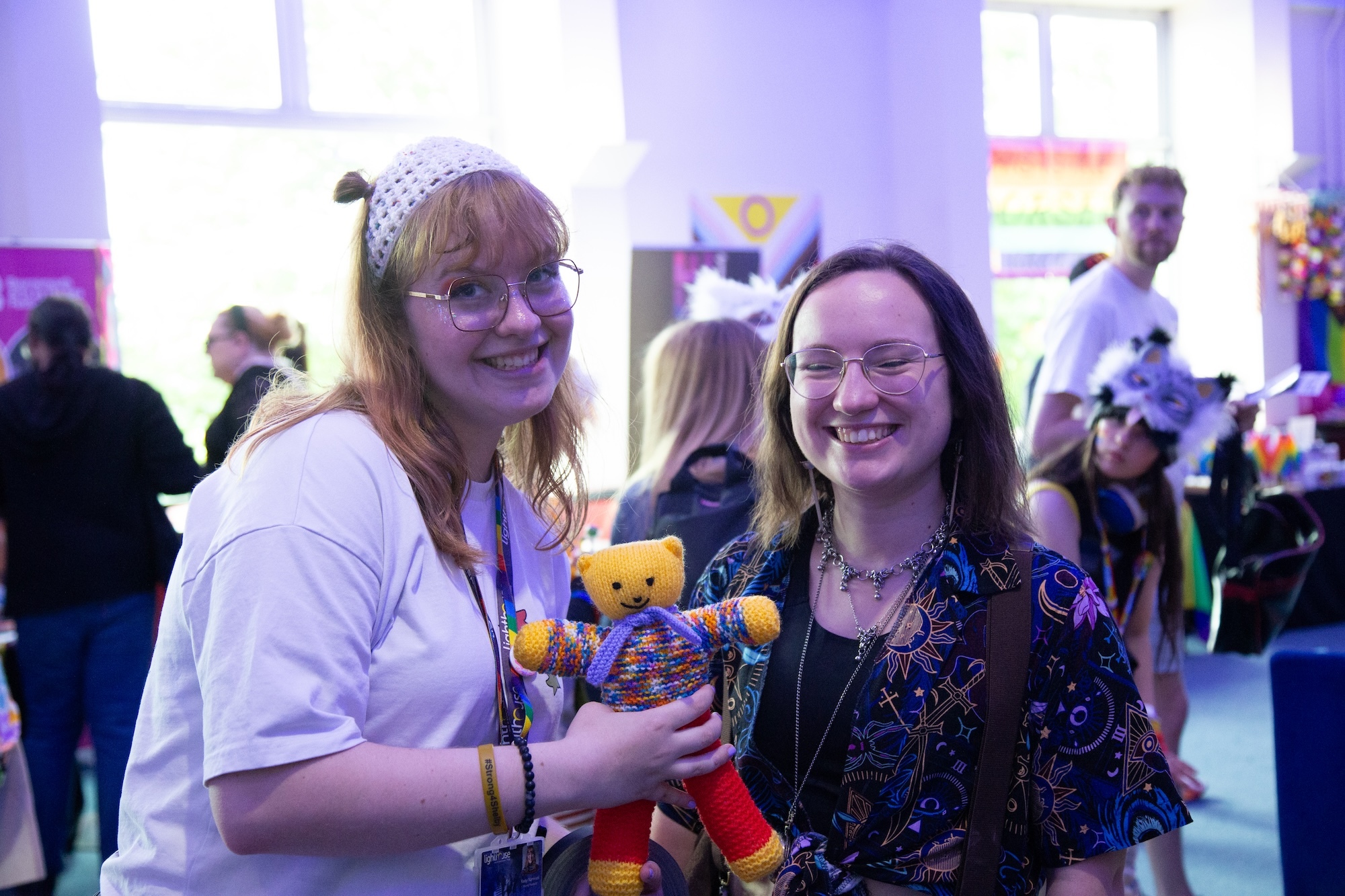 Two young people smiling at camera, one holding small teddy bear, standing in crowded purple-lit room with windows in background.