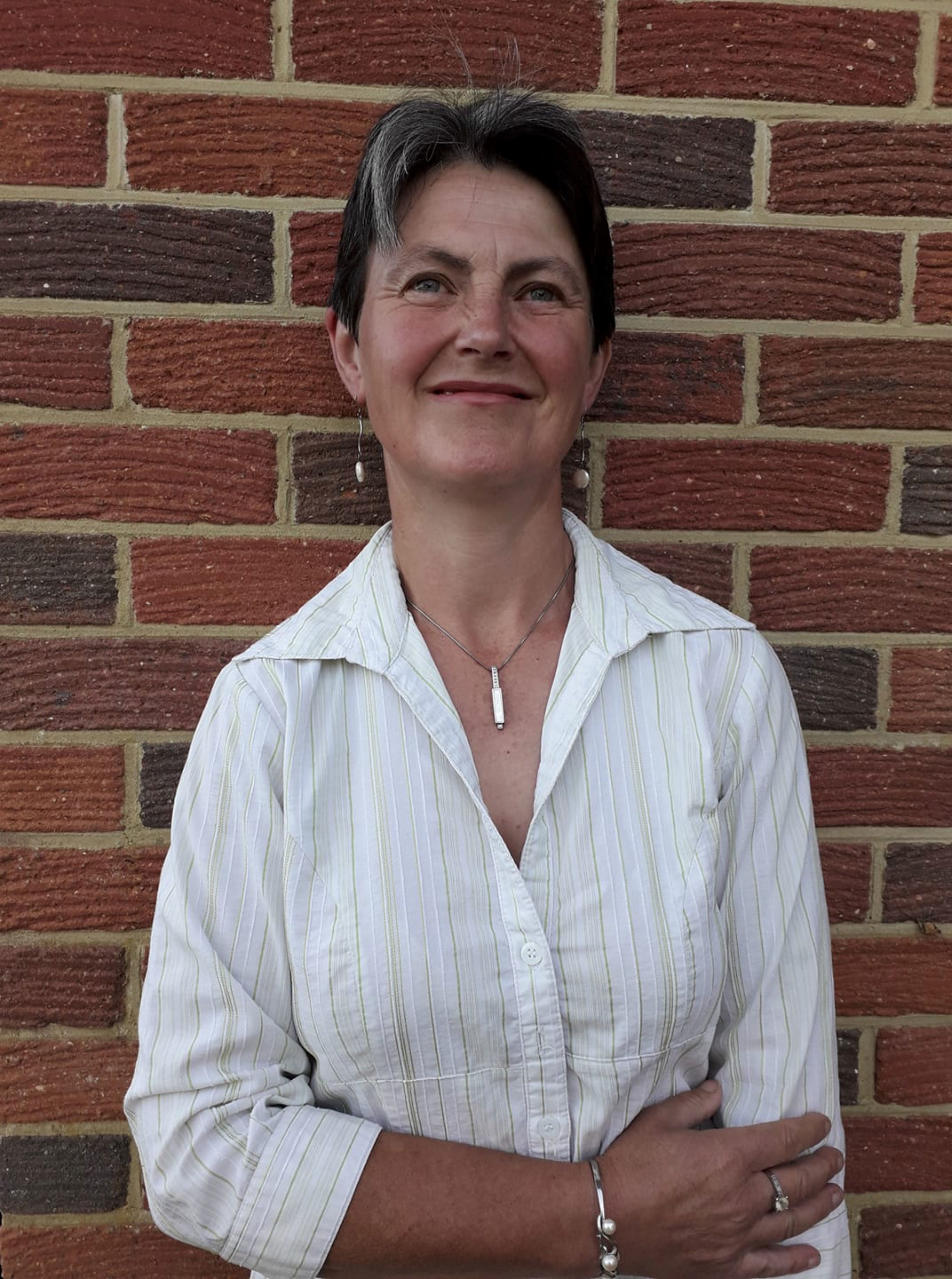 Photo of Kate Couch against a brick wall, smiling and wearing a white shirt, necklace and bracelet. She has short dark-grey hair.
