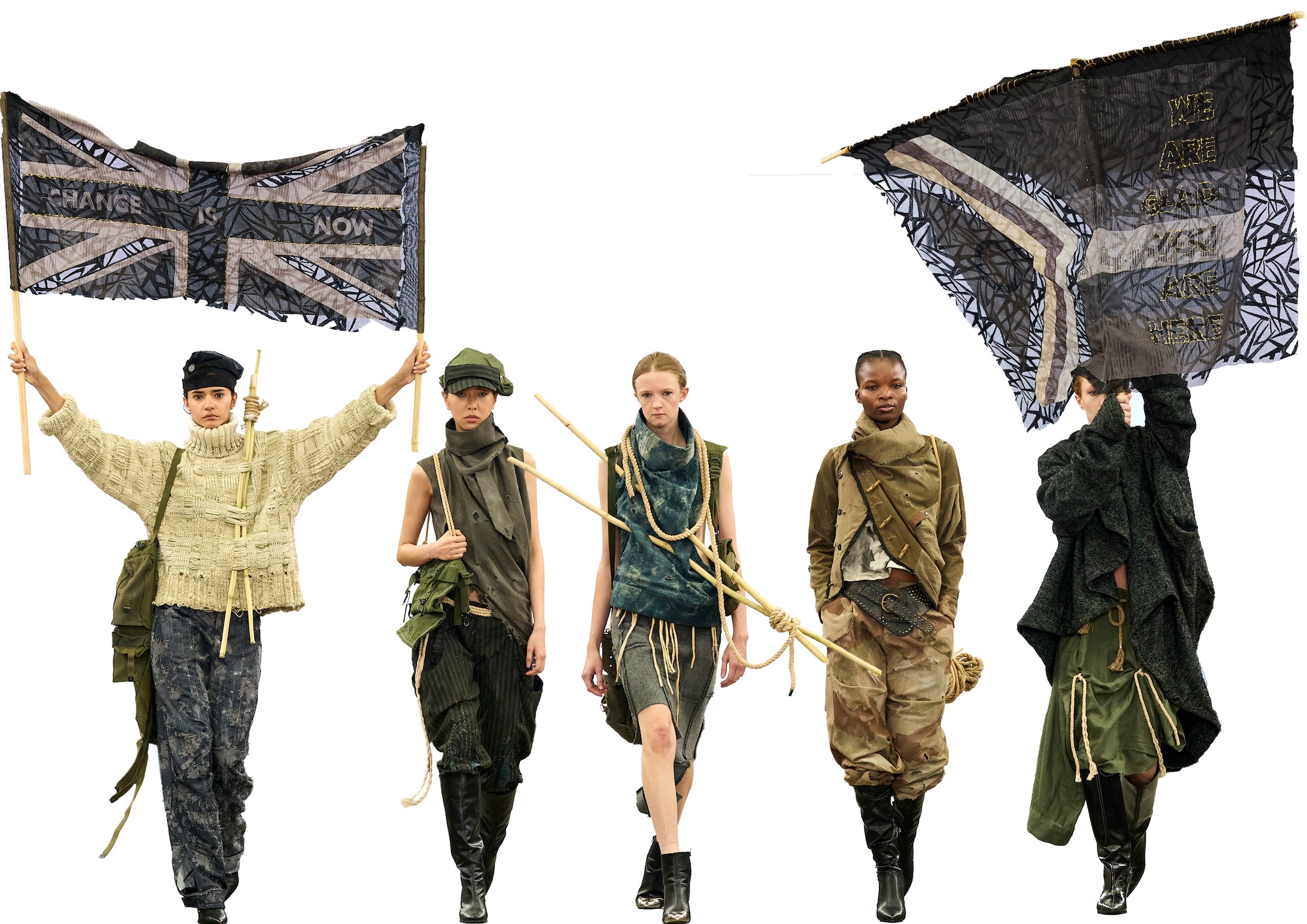 Five fashion models in layered outfits walk against a white background, with two carrying large black patchwork flags.