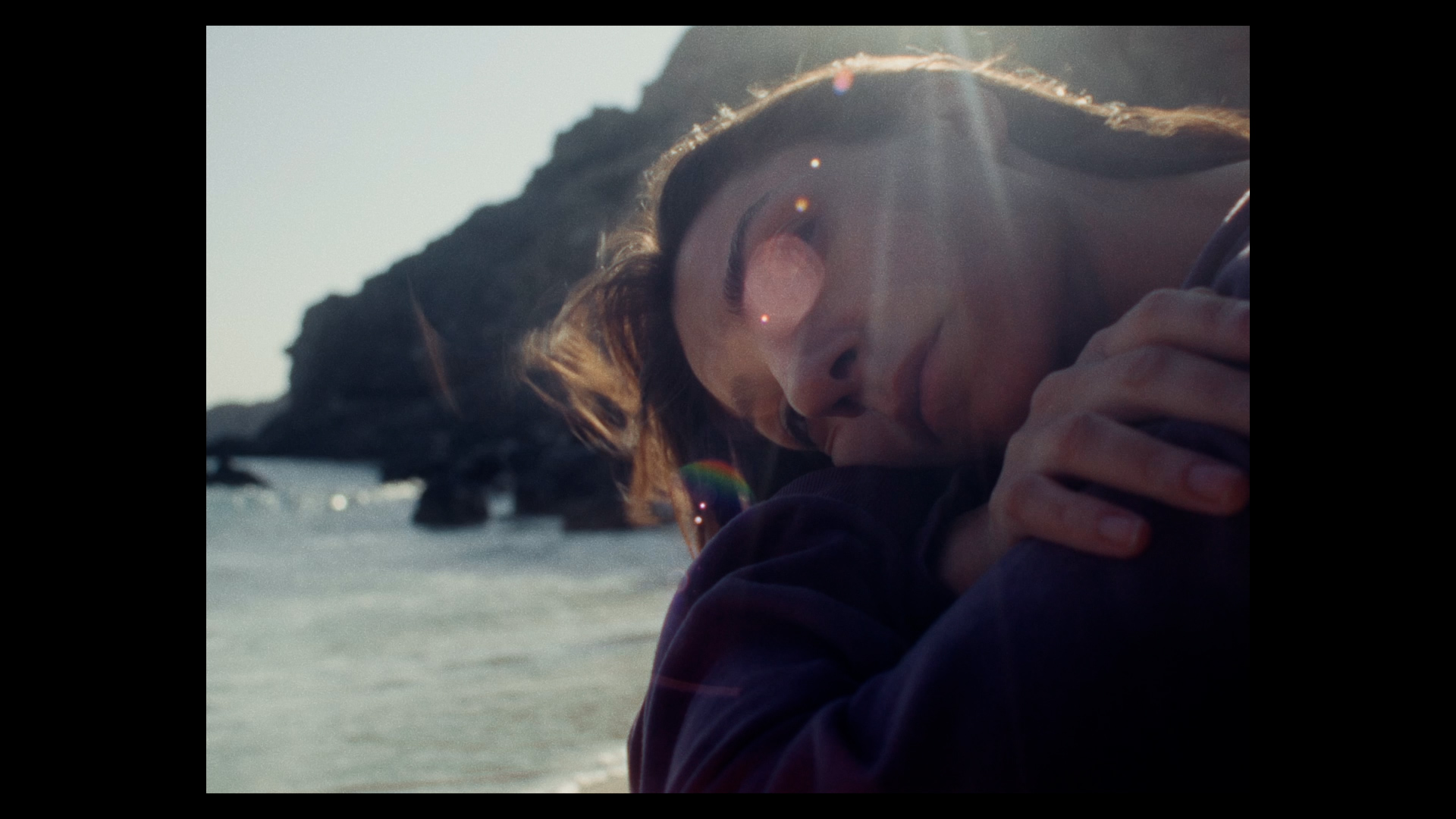 Close-up film still of a person hugging themselves looking sad with a shoreline in the background.