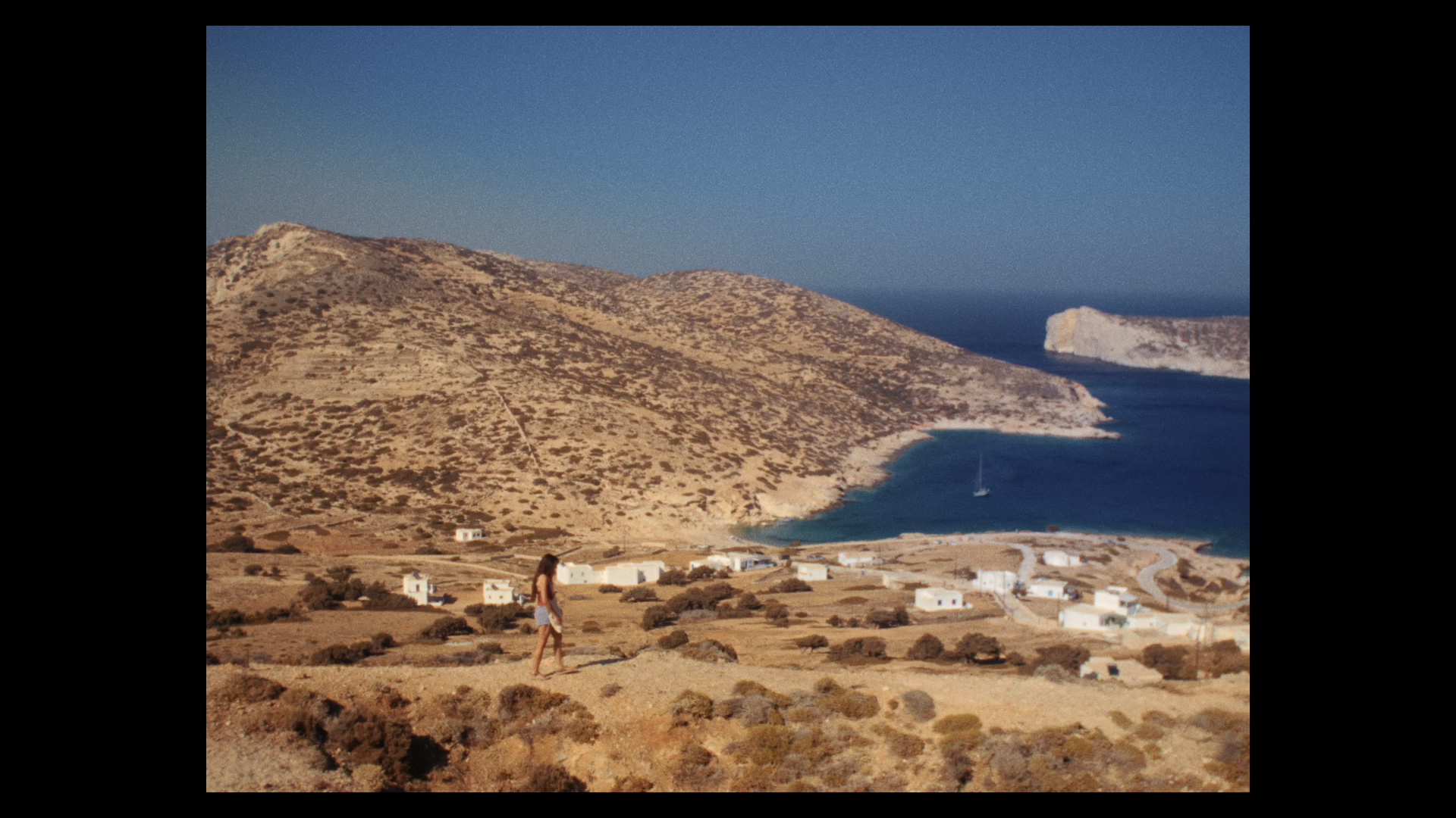 Film still of a person walking across a gravel path with an Greek hill and mountain vista in the background.