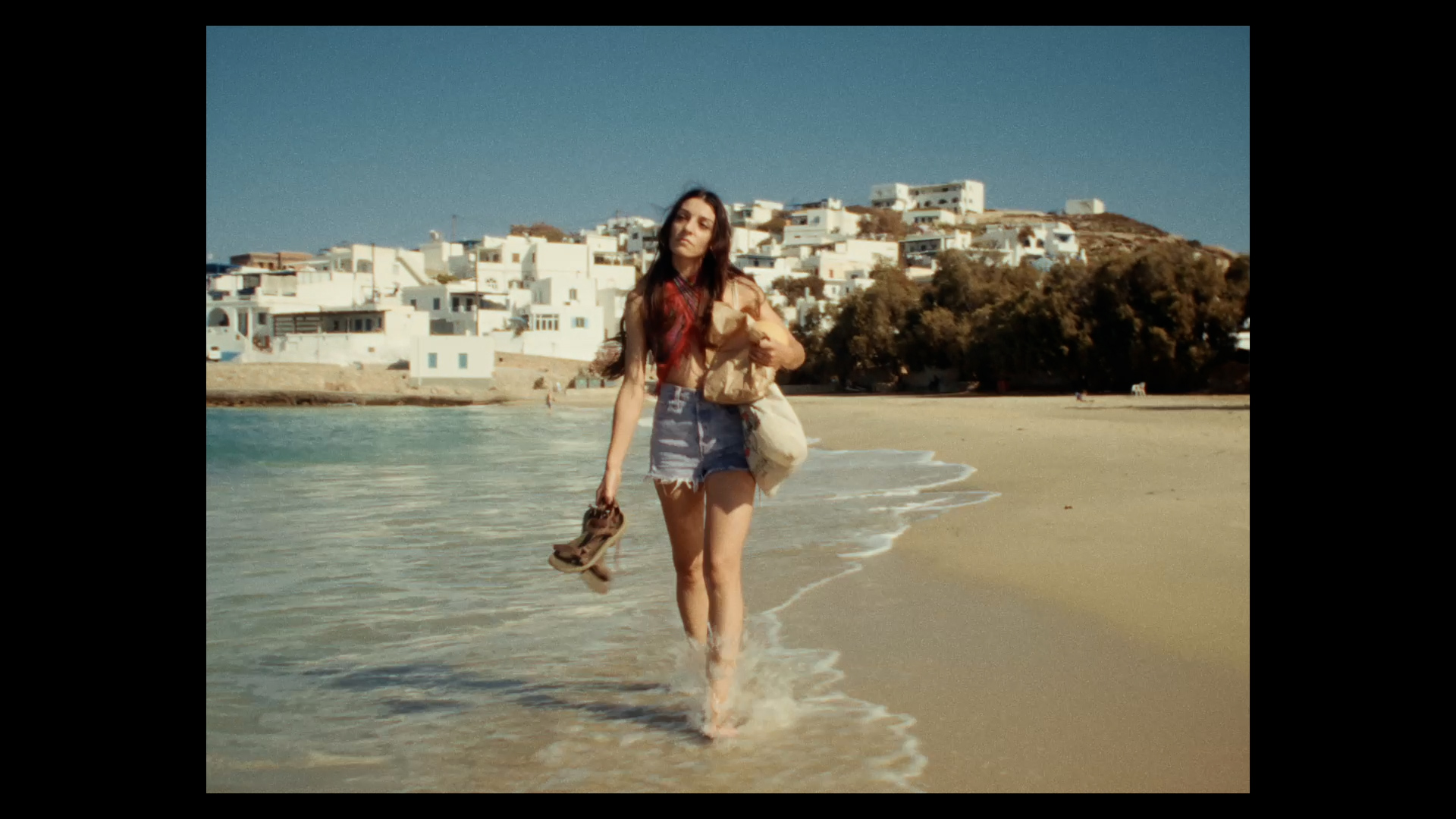 Film still of a young person carrying their shoes in one hand and a bag on their shoulder walking through water on a beach shoreline.