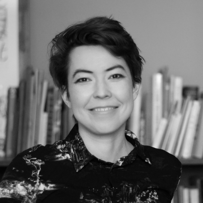 Black and white portrait of woman with short dark hair wearing patterned dark top, smiling at camera with bookshelves behind her.