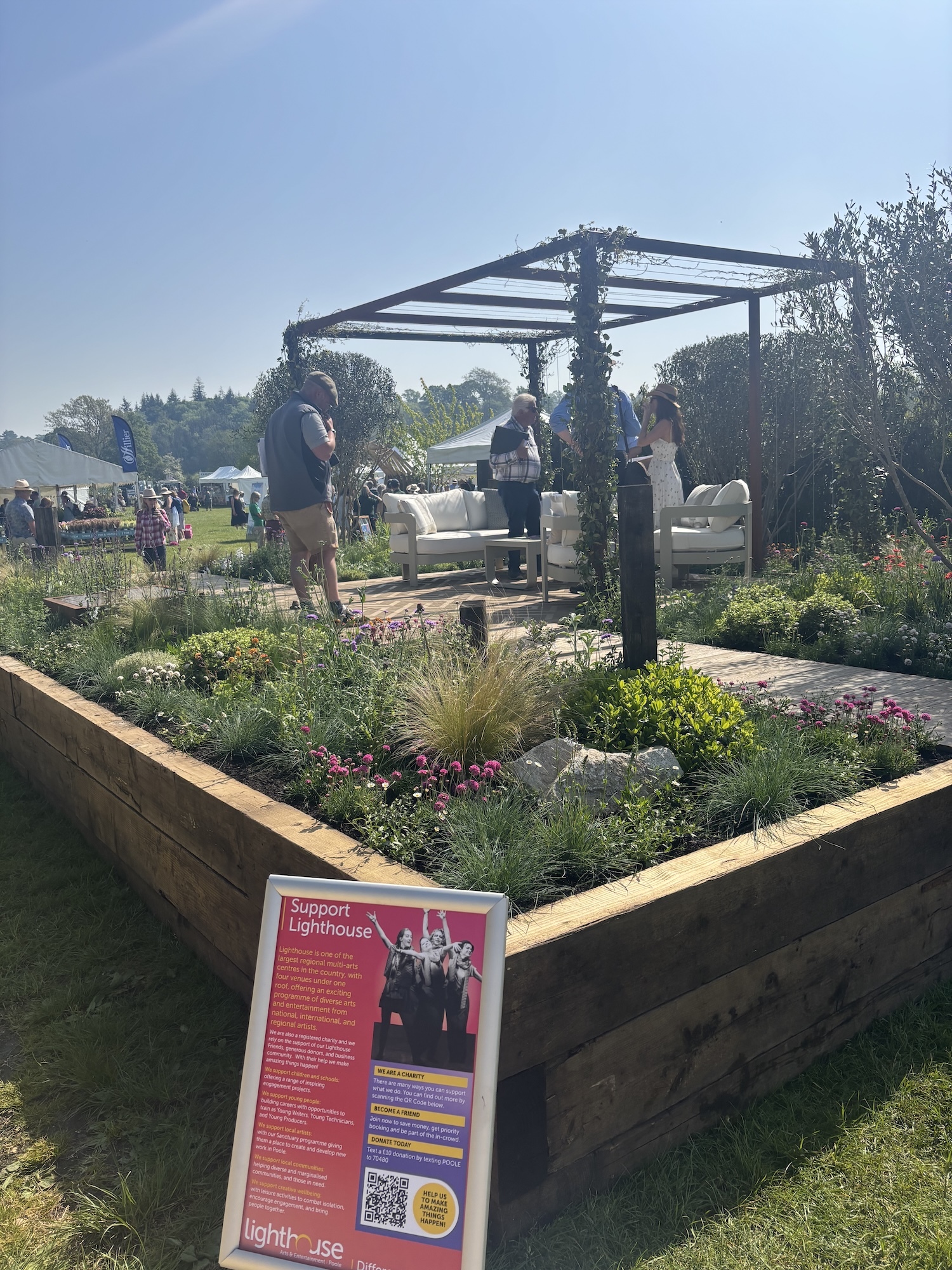 Garden display with raised wooden beds, metal pergola, outdoor seating area and red charity sign. People viewing under blue sky.