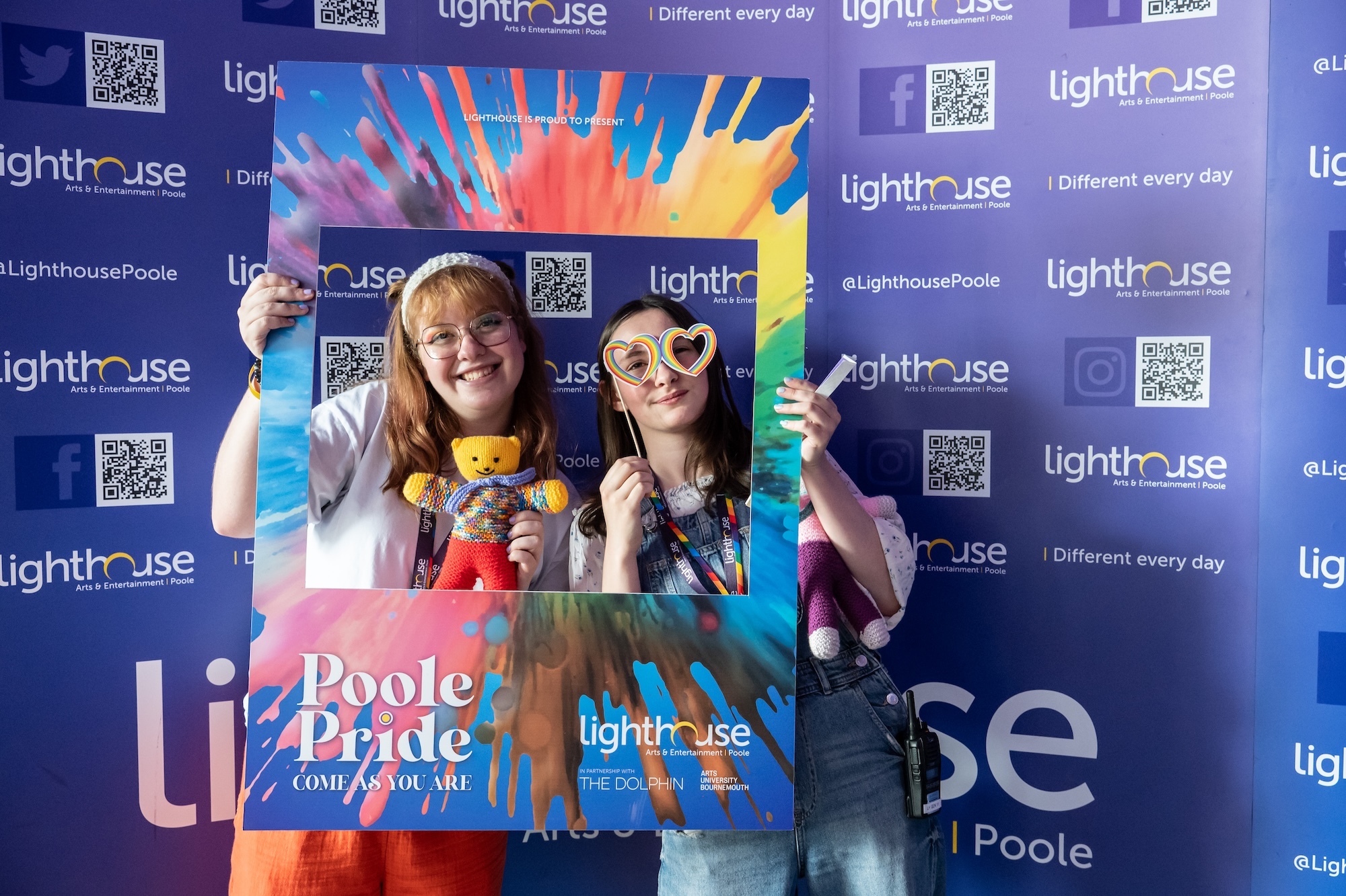 Two people holding colourful cardboard frame at Lighthouse Poole event, blue branded backdrop with QR codes, rainbow burst design on frame