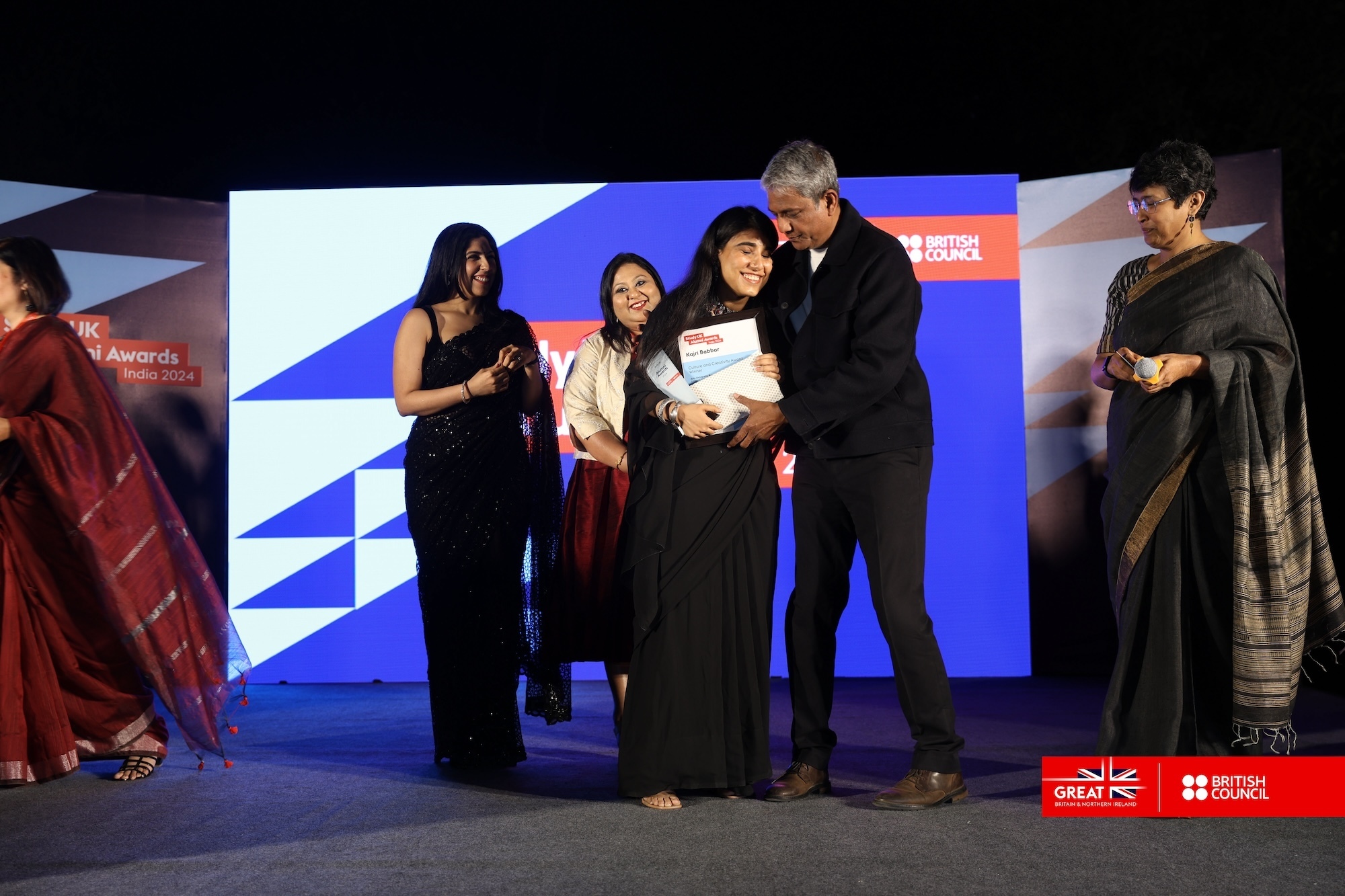 K Babbar on stage with other British Council Study UK Alumni Awards as she receives her award, a blue and white abstract panel is in the background.