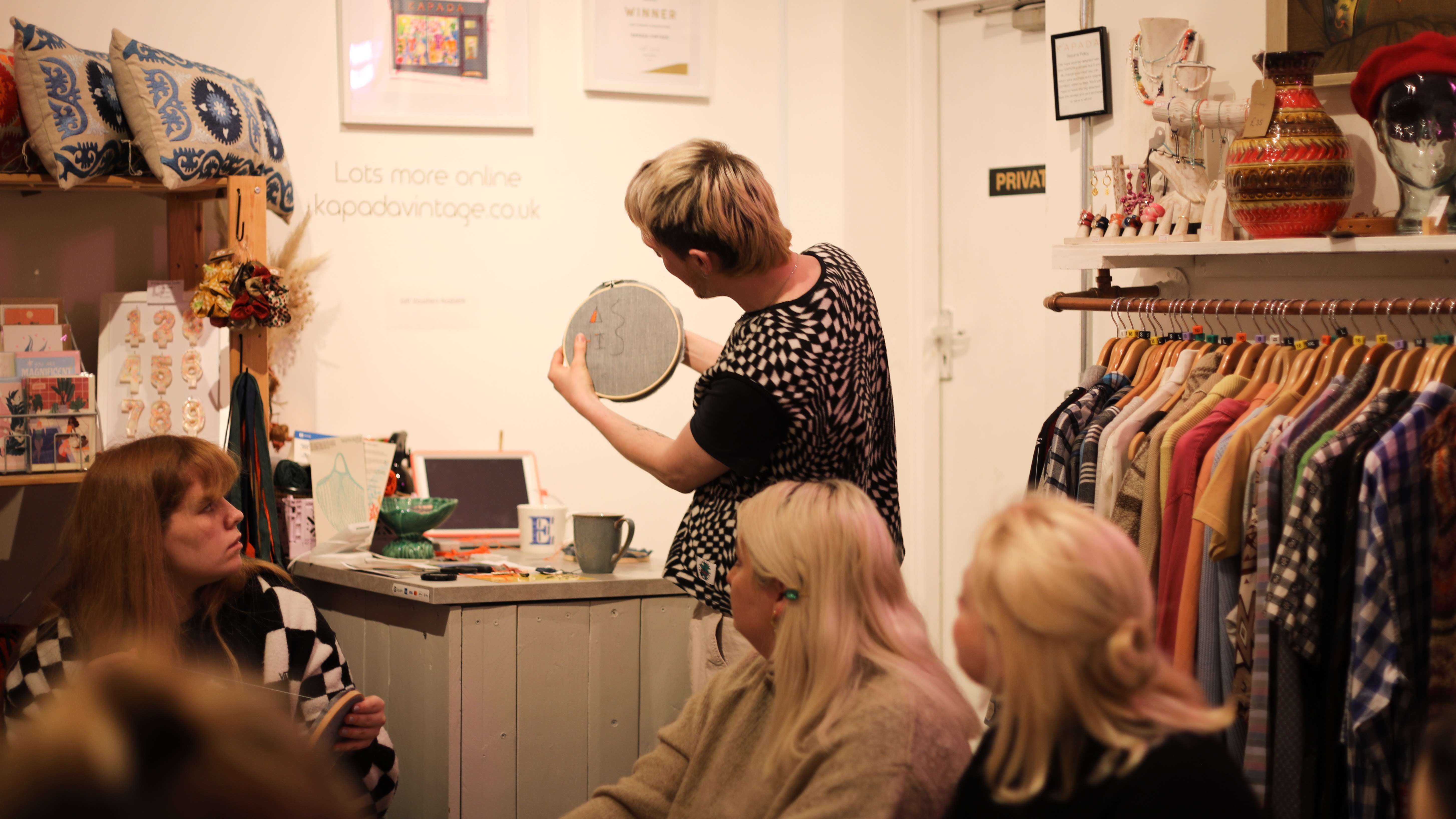 A person stands in front of a small class of people and demonstrates how to embroider.
