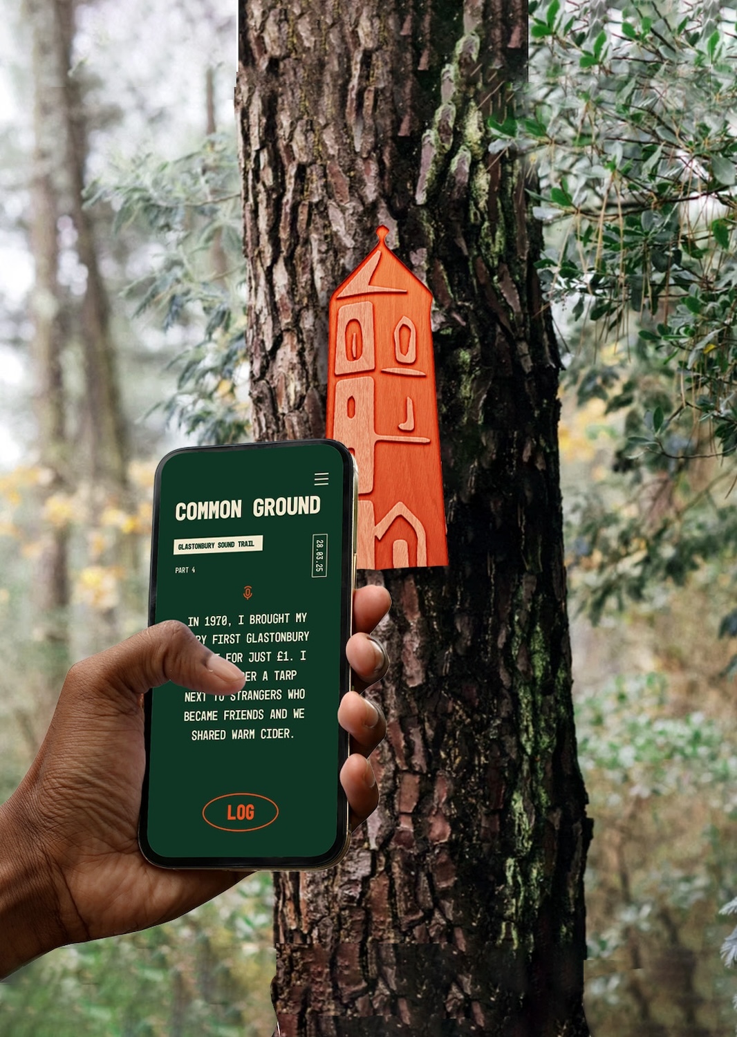 Hand holding green smartphone displaying "Common Ground" text beside orange decorative house ornament hanging on tree trunk in forest.