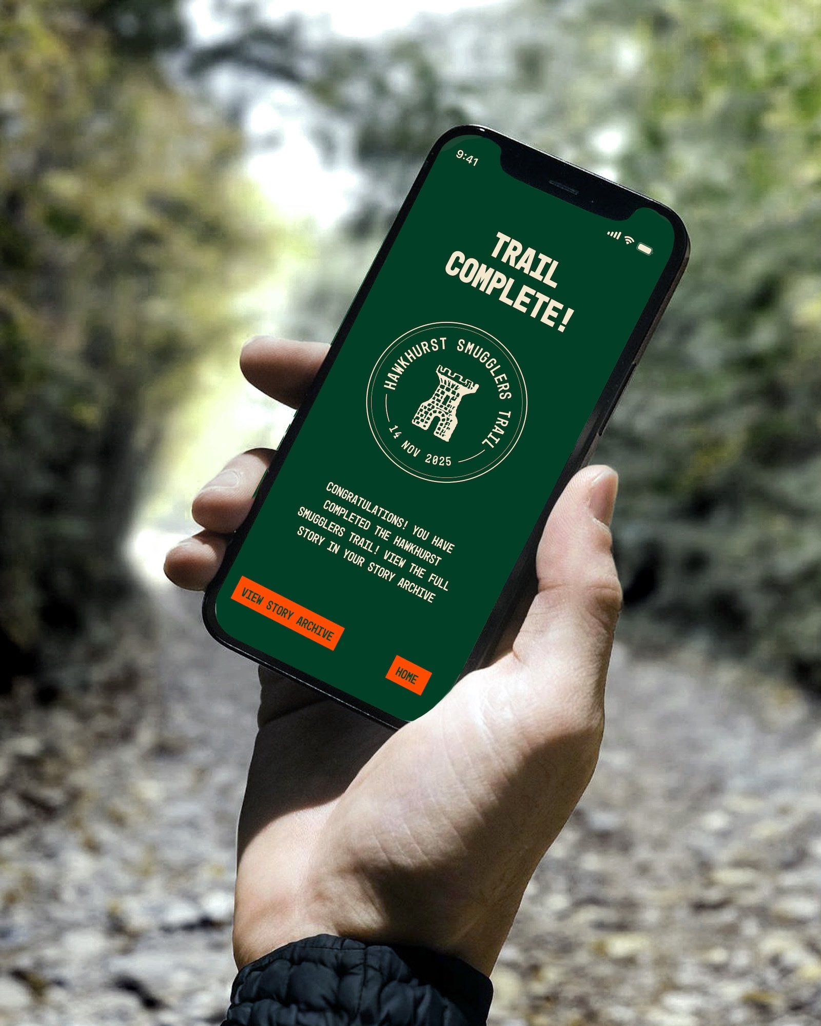 Hand holding smartphone displaying green screen with "TRAIL COMPLETE!" text, circular badge with tree icon, and orange button, outdoors.