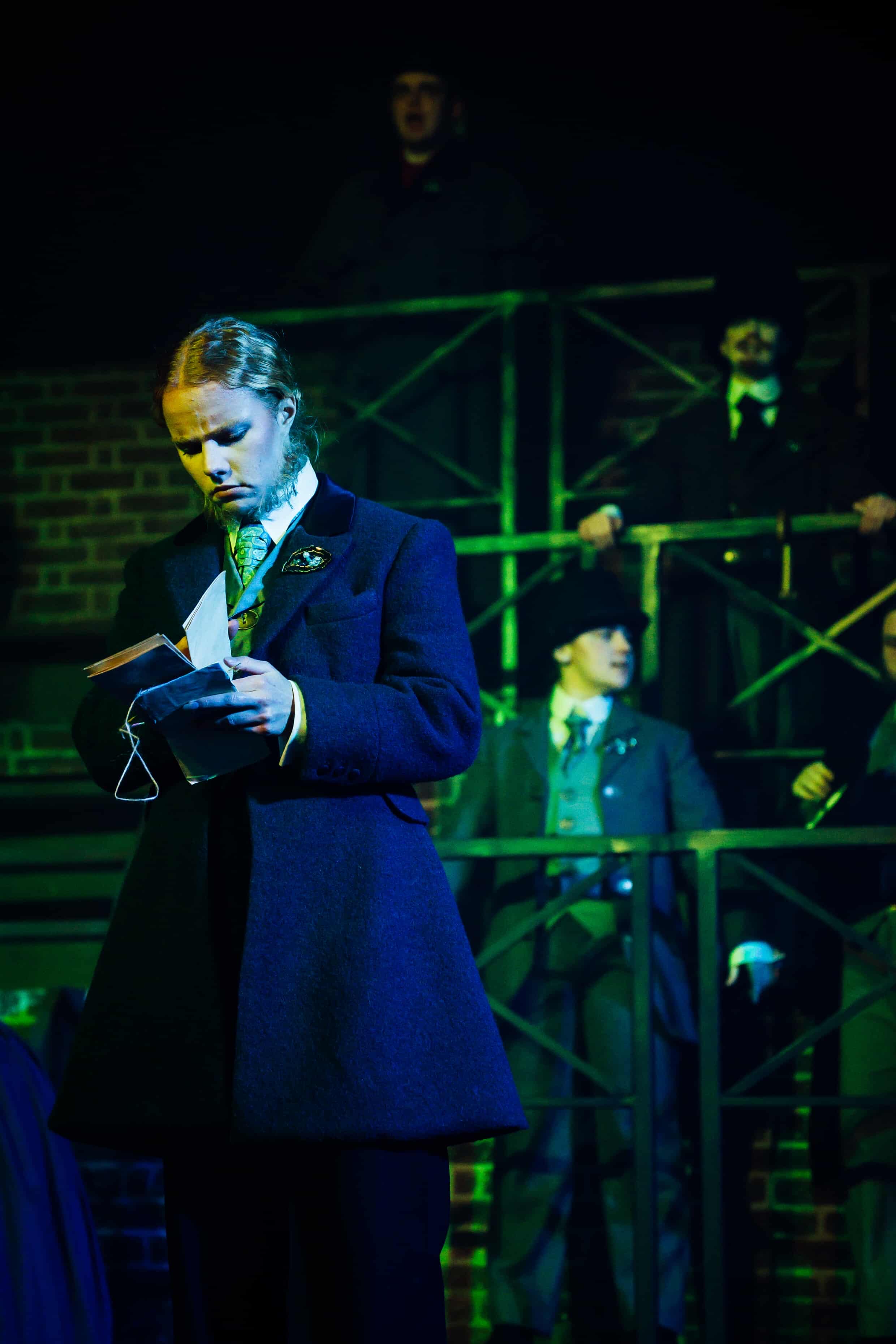 A man dressed in a black coat and waistcoat reads from a letter while standing in a spotlight. In the background, several figures in Victorian attire observe from a raised platform against a darkened stage set.