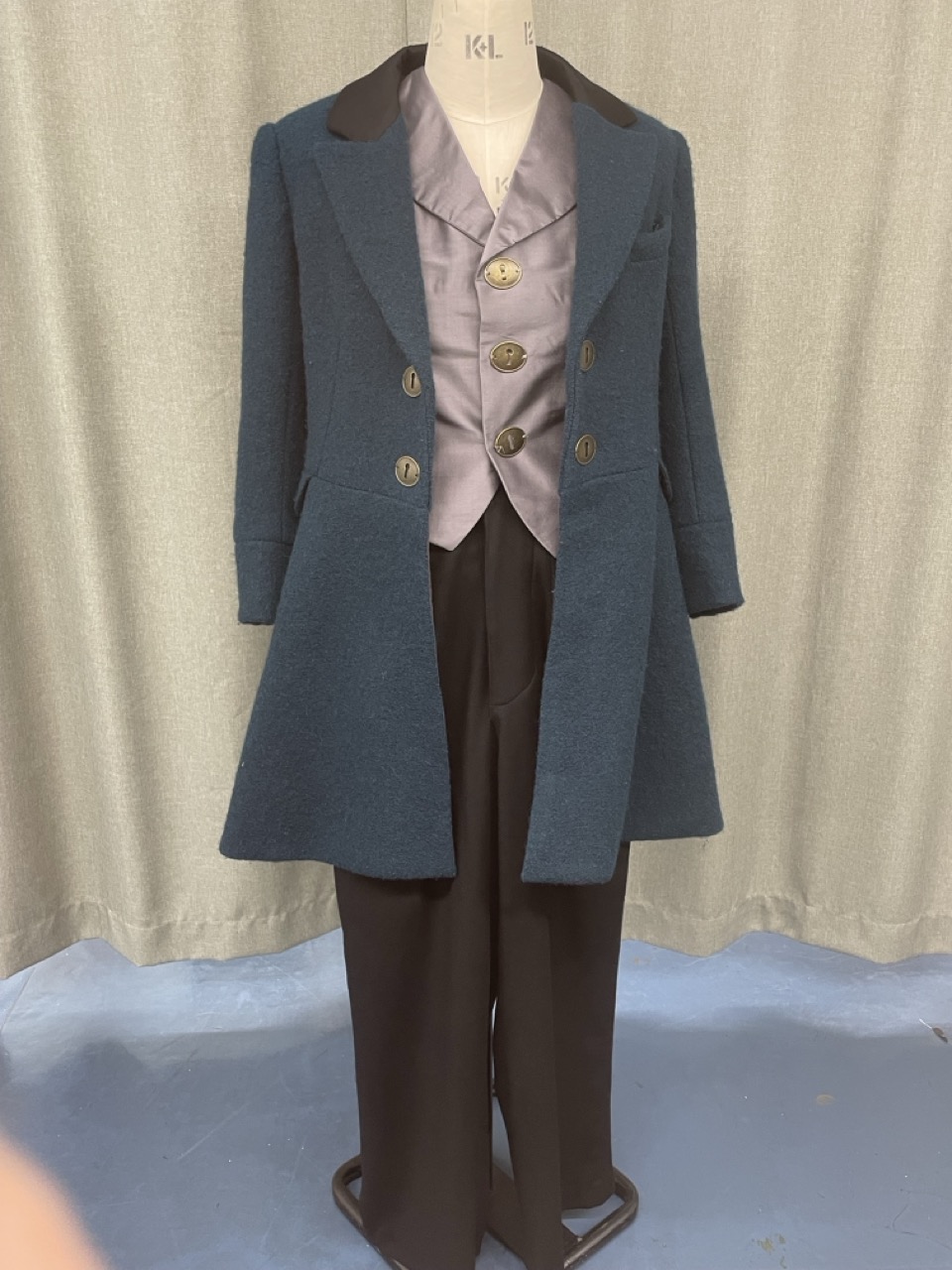 A vintage-style outfit displayed on a dress form, featuring a dark blue coat with black lapels, a light grey shirt, and tailored black trousers. The coat has large buttons and a tailored fit, creating a classic appearance.