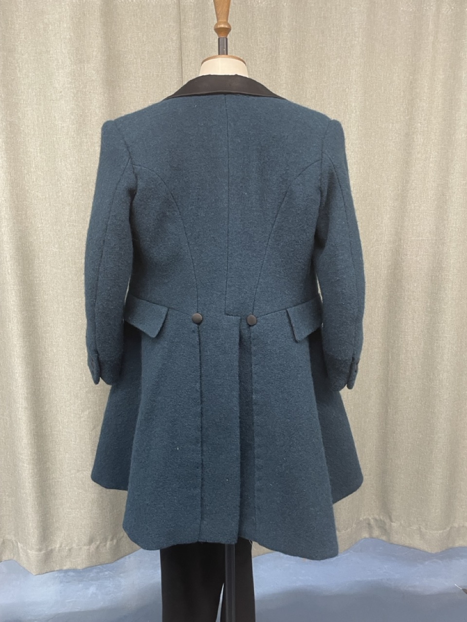 A dark blue wool coat displayed on a mannequin, featuring a black collar and a flared design. The back of the coat shows two buttons at the waist level, with a plain, unembellished fabric draping below.