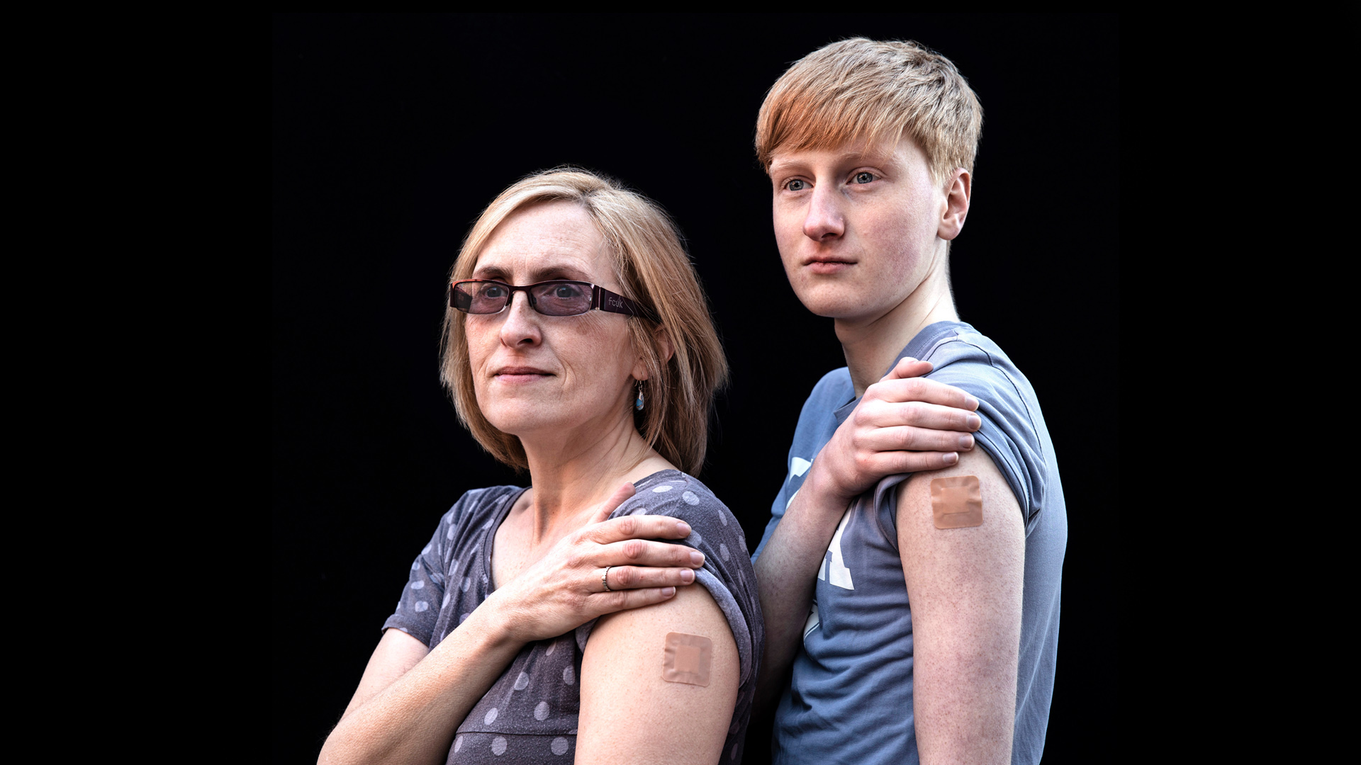Two people stand with their right hands pulling up their left sleeves and showing square tattoos on their upper arms.