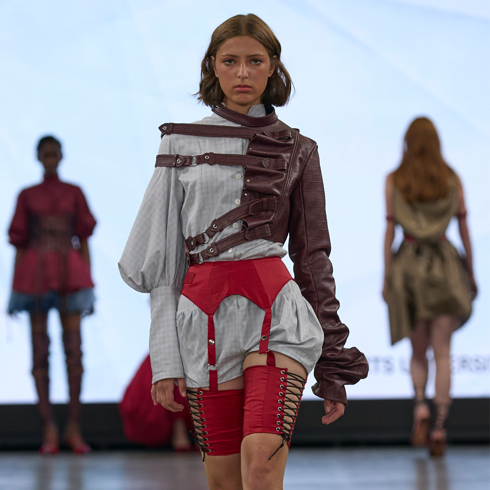 Model walks runway wearing white blouse with brown leather harness-style straps and red lace-up shorts, two blurred figures in background.