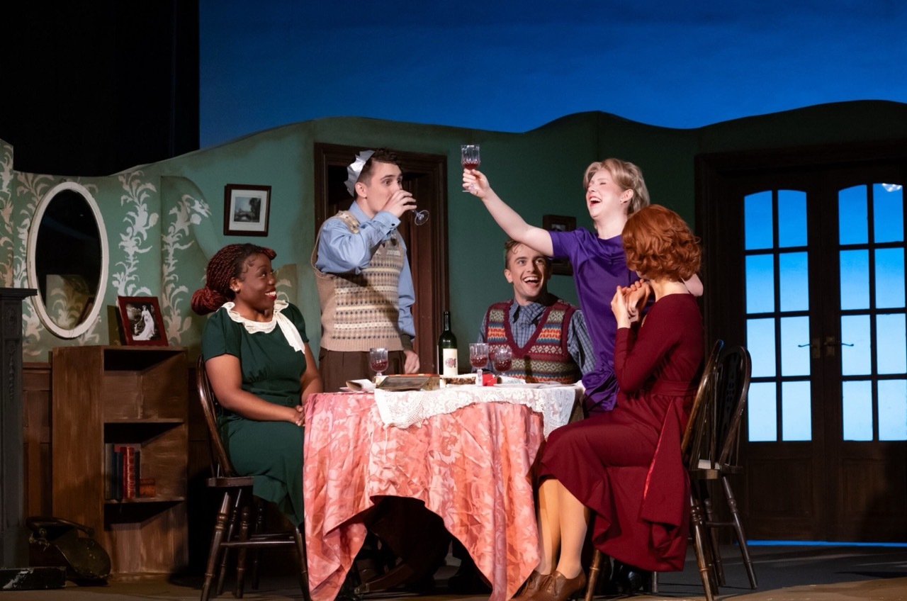 Five actors on stage around dining table with pink tablecloth. Green wallpaper, oval mirror, large windows show blue sky and hills.