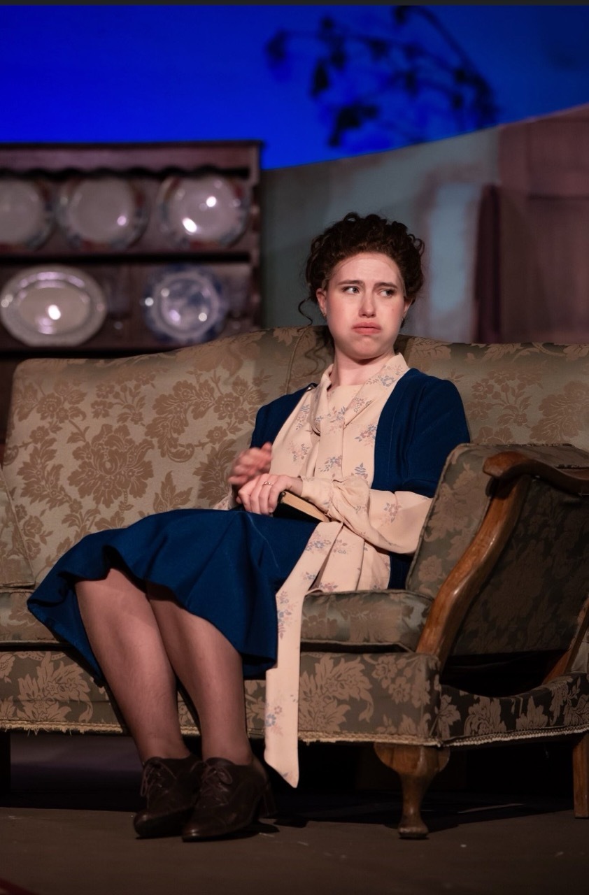 Woman in period costume with dark blue dress and cream waistcoat seated on ornate sofa, kitchen dresser with plates visible behind.