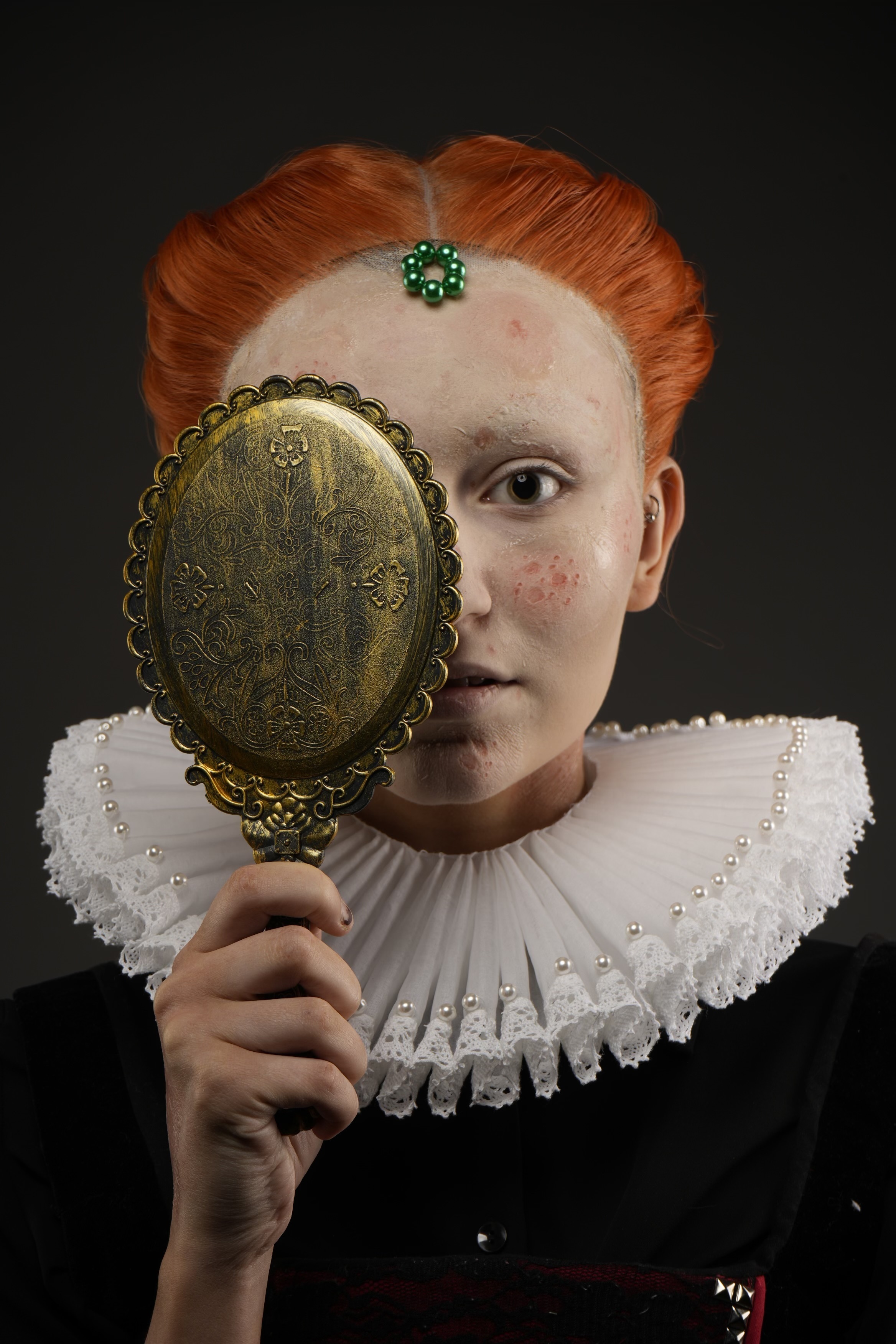Woman with red hair and white ruff collar holds ornate brass hand mirror, green jewel on forehead, dark background.