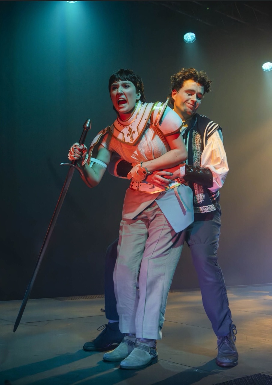 Two male performers on stage under coloured lights, one with fake blood makeup holding a microphone stand, the other embracing him from behind.