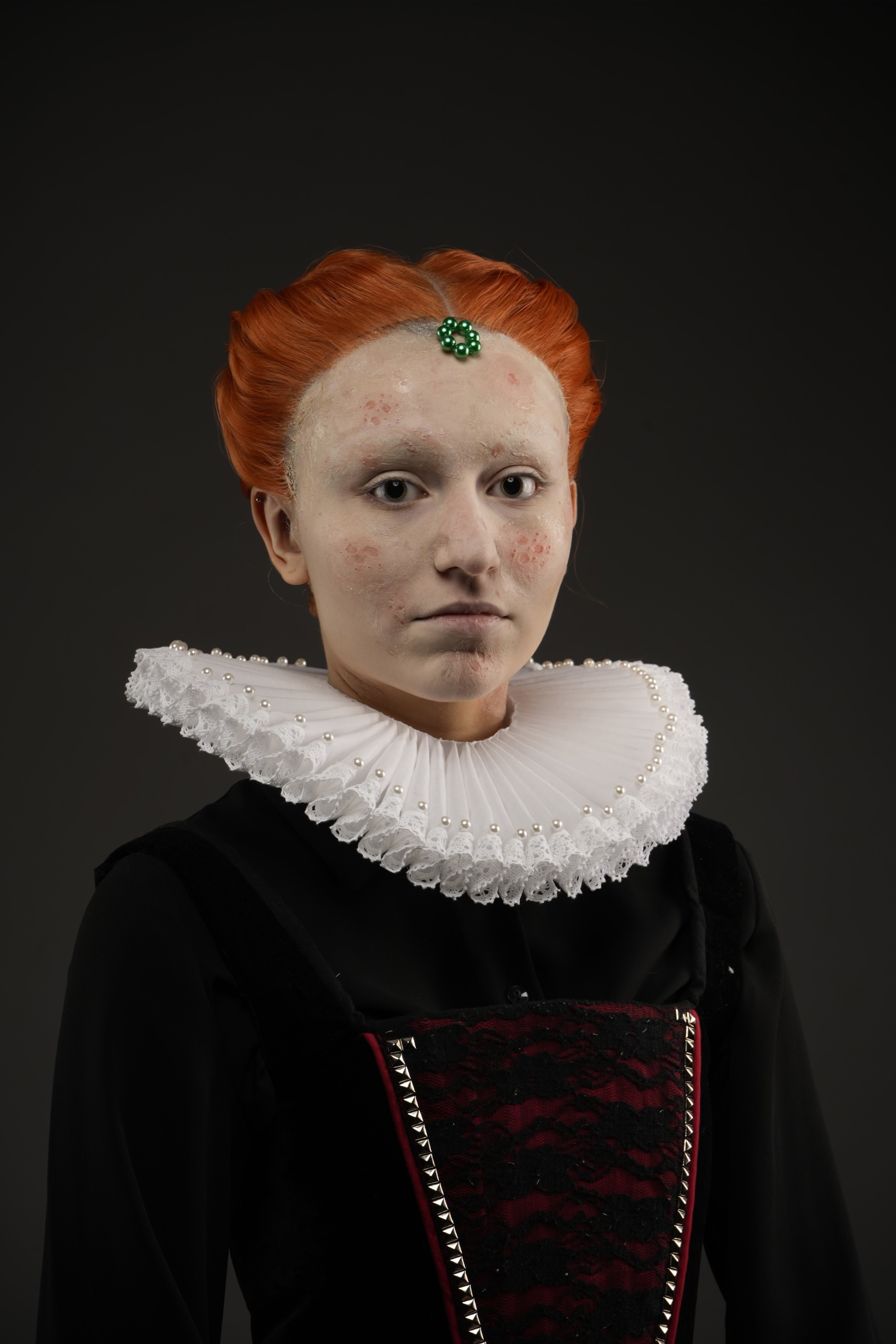 Woman with orange hair and green forehead jewel wearing white ruffled collar and black dress with red bodice against dark background.