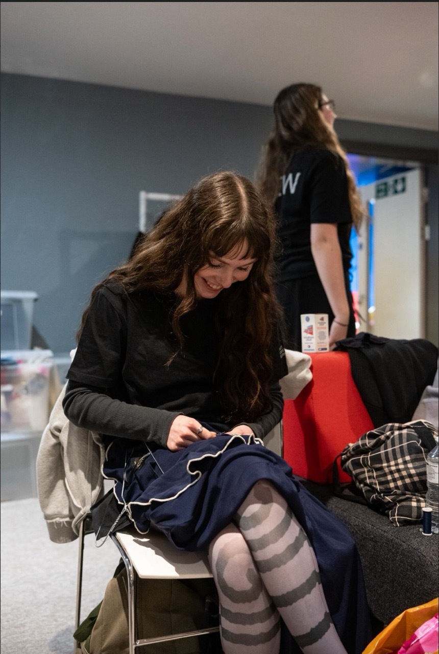 Woman in black coat sitting with striped tights, another person standing behind her in indoor setting with bags and belongings.