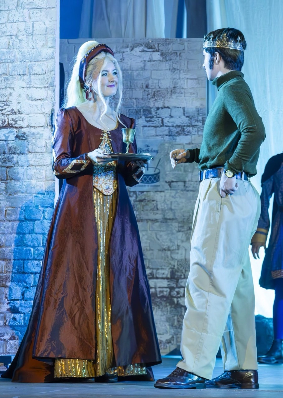 Two performers in period costume on stage; woman in burgundy medieval dress with white headdress, man in green jacket with crown.