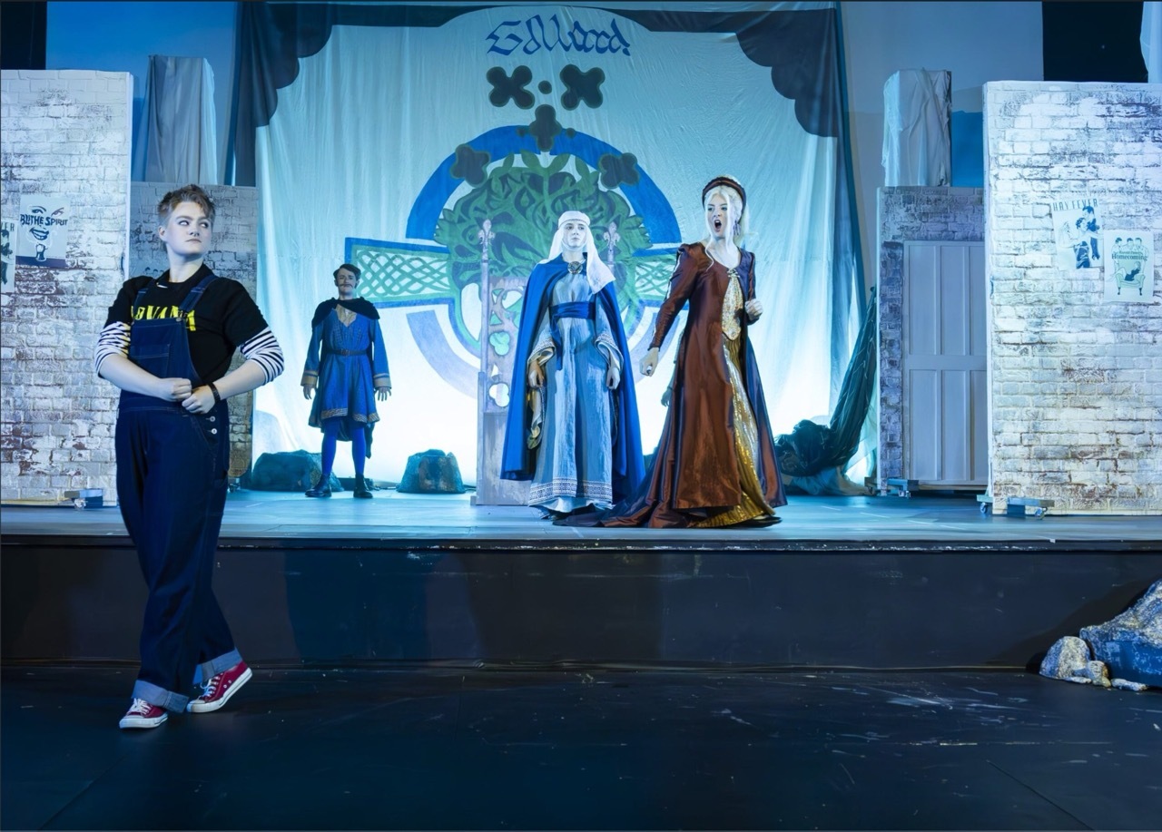Theatre stage with medieval-costumed performers in blue and burgundy robes, stone column backdrop, person in black tracksuit standing in foreground.