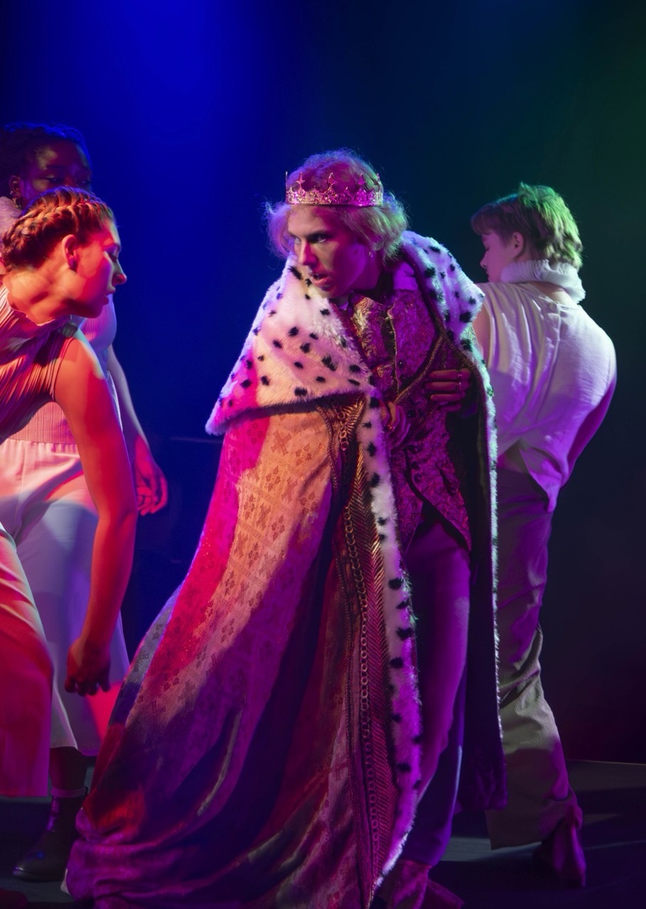 Three actors on stage under blue and purple lighting. Centre figure wears ornate robes and crown, flanked by two others in period costume.