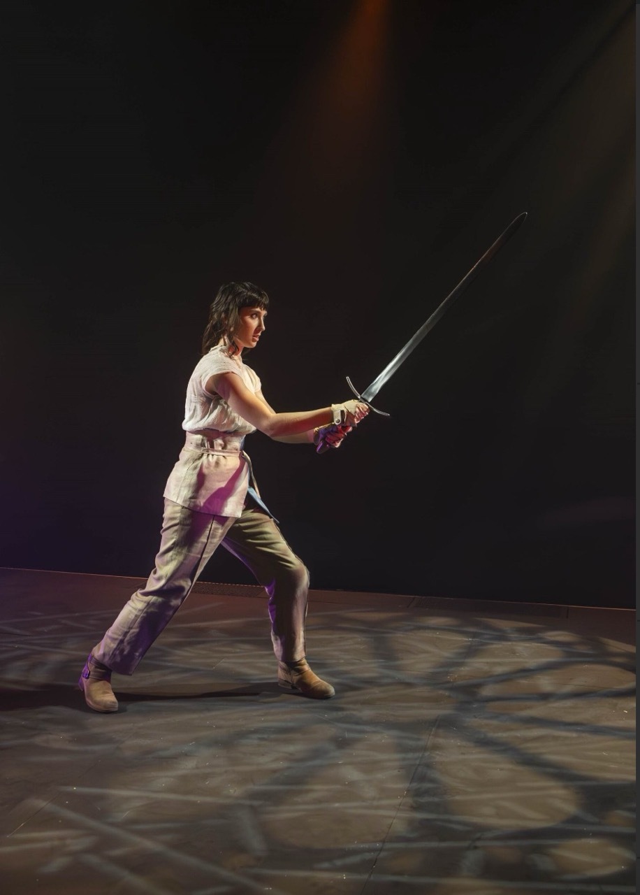 Person in white costume with purple sash holds sword in lunging position on tiled floor against dark background with coloured lights.