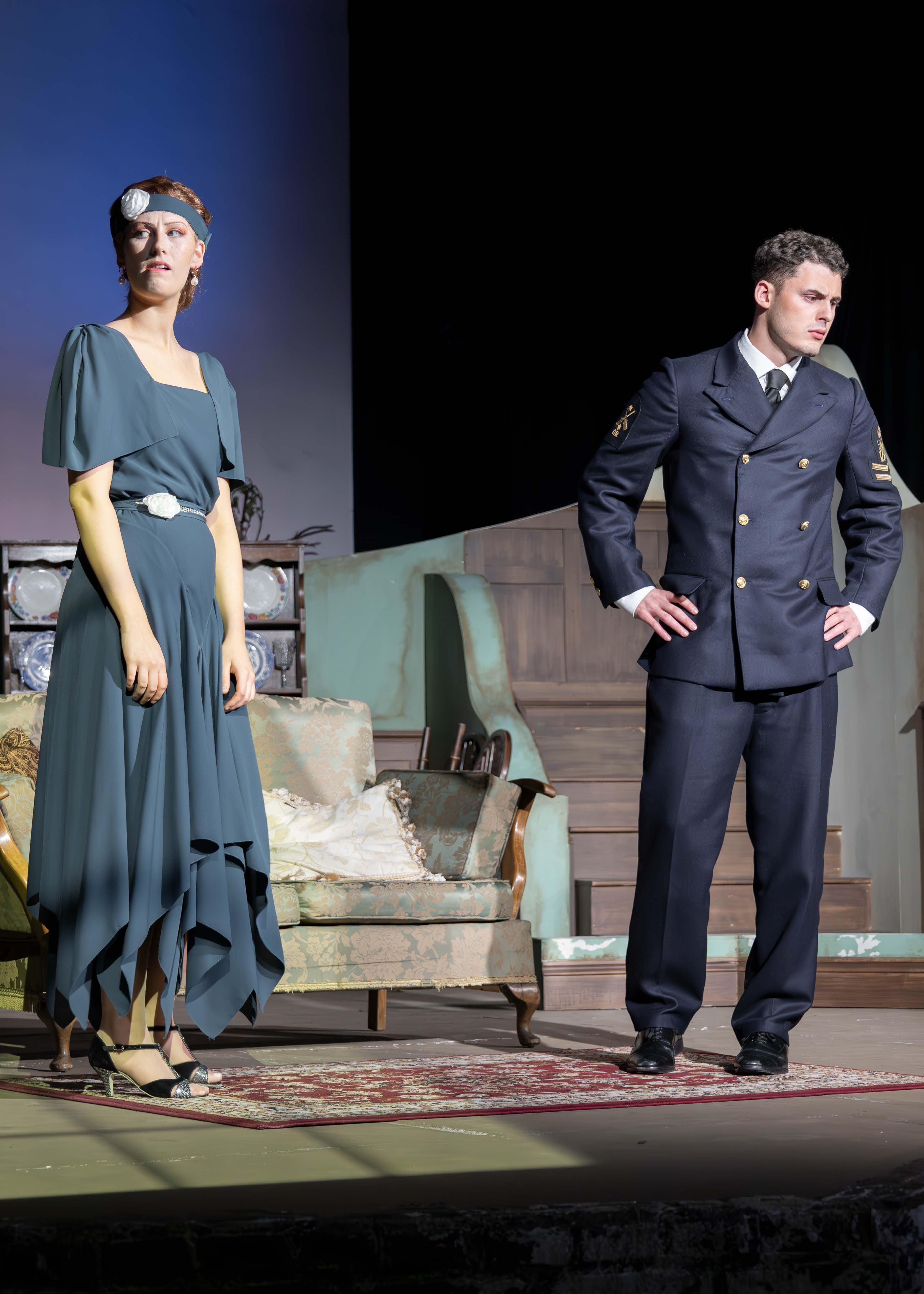 Two BA (Hons) Acting students on stage: one in teal 1930s dress with headband, the other in dark military uniform. Theatre set with sofa and stone wall backdrop.