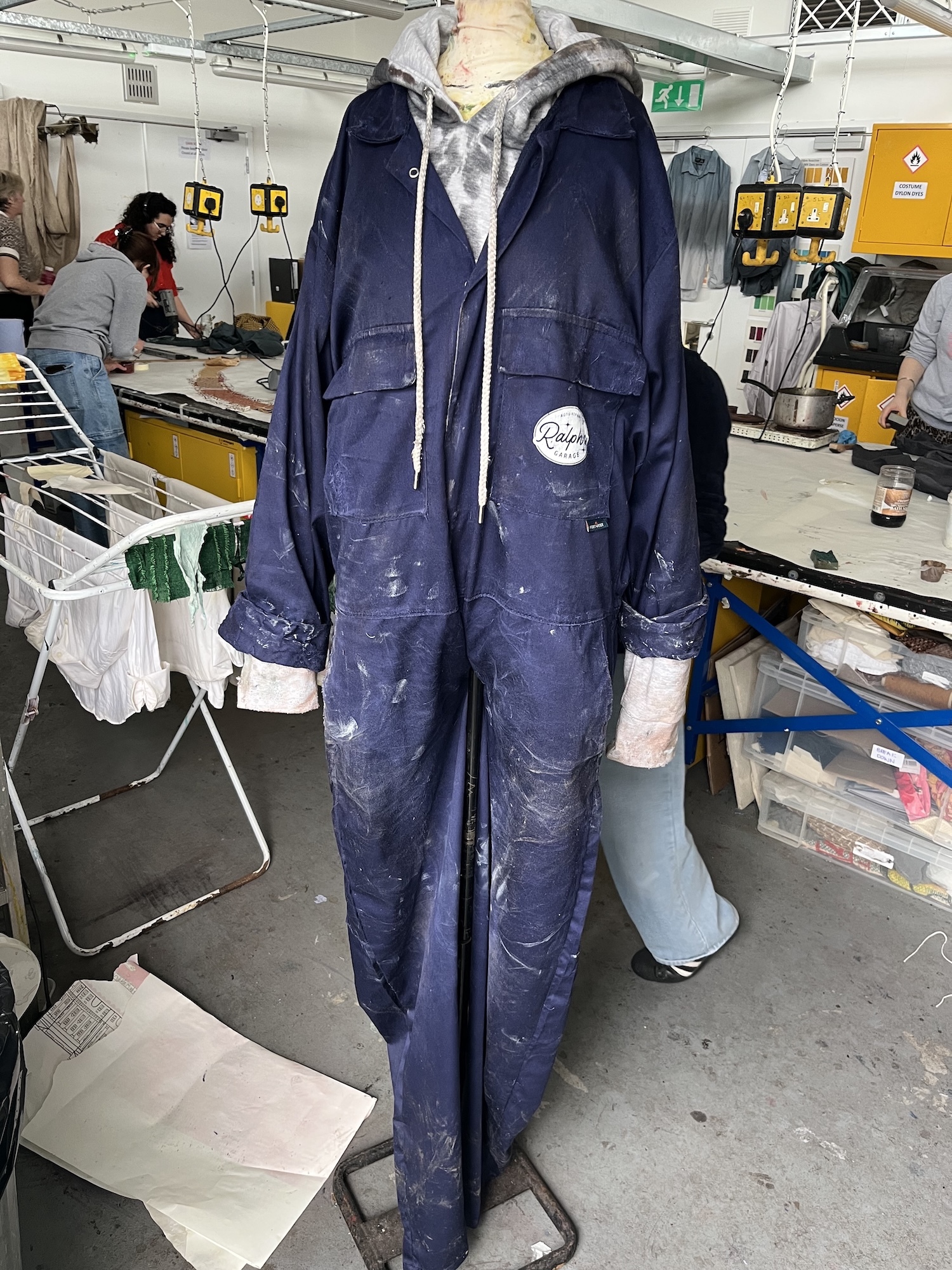 Navy blue coveralls with grey hood hanging in ship's workspace. People working at tables with yellow equipment in background.