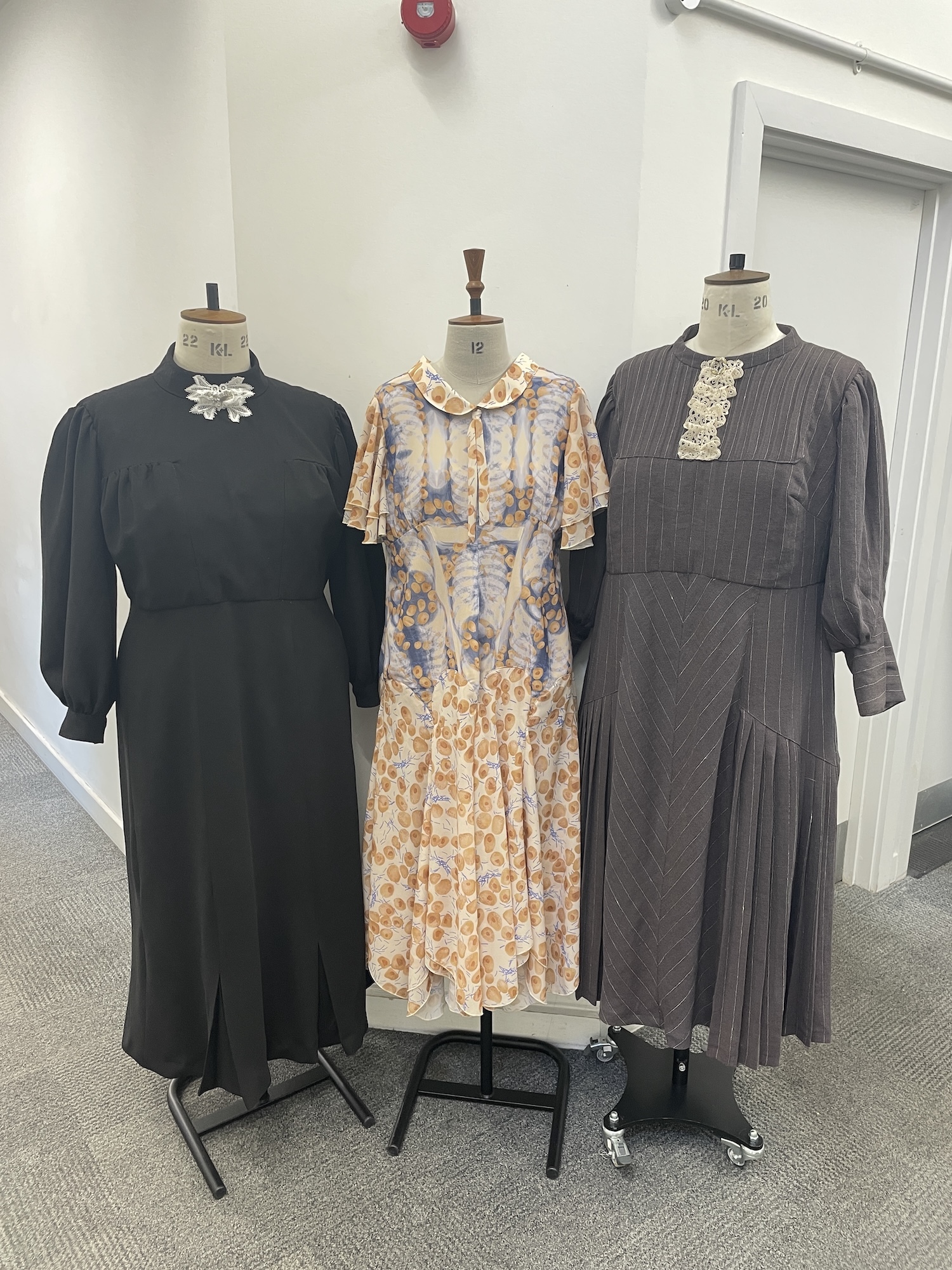 Three vintage dresses on mannequins: dark green with white lace collar, beige floral print with ruffles, dark grey with lace trim.