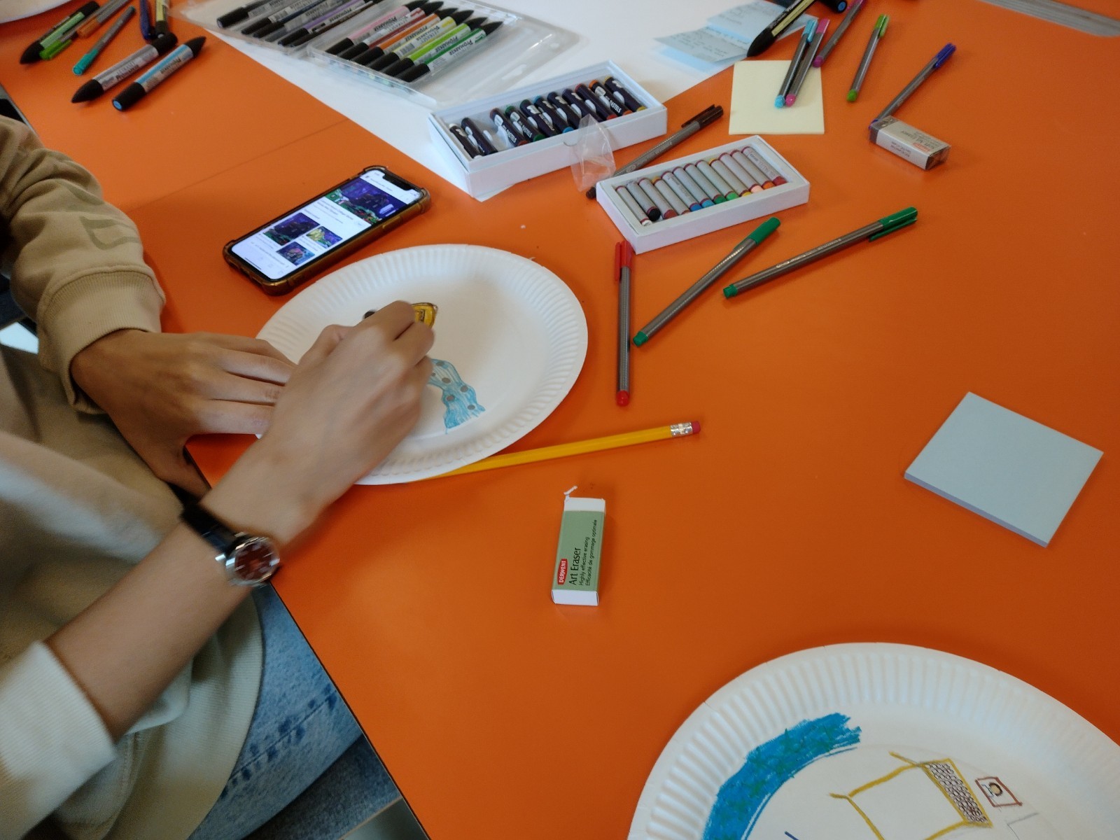 Someone sitting at a table and drawing on a paper plate, with art supplies scattered around the table.