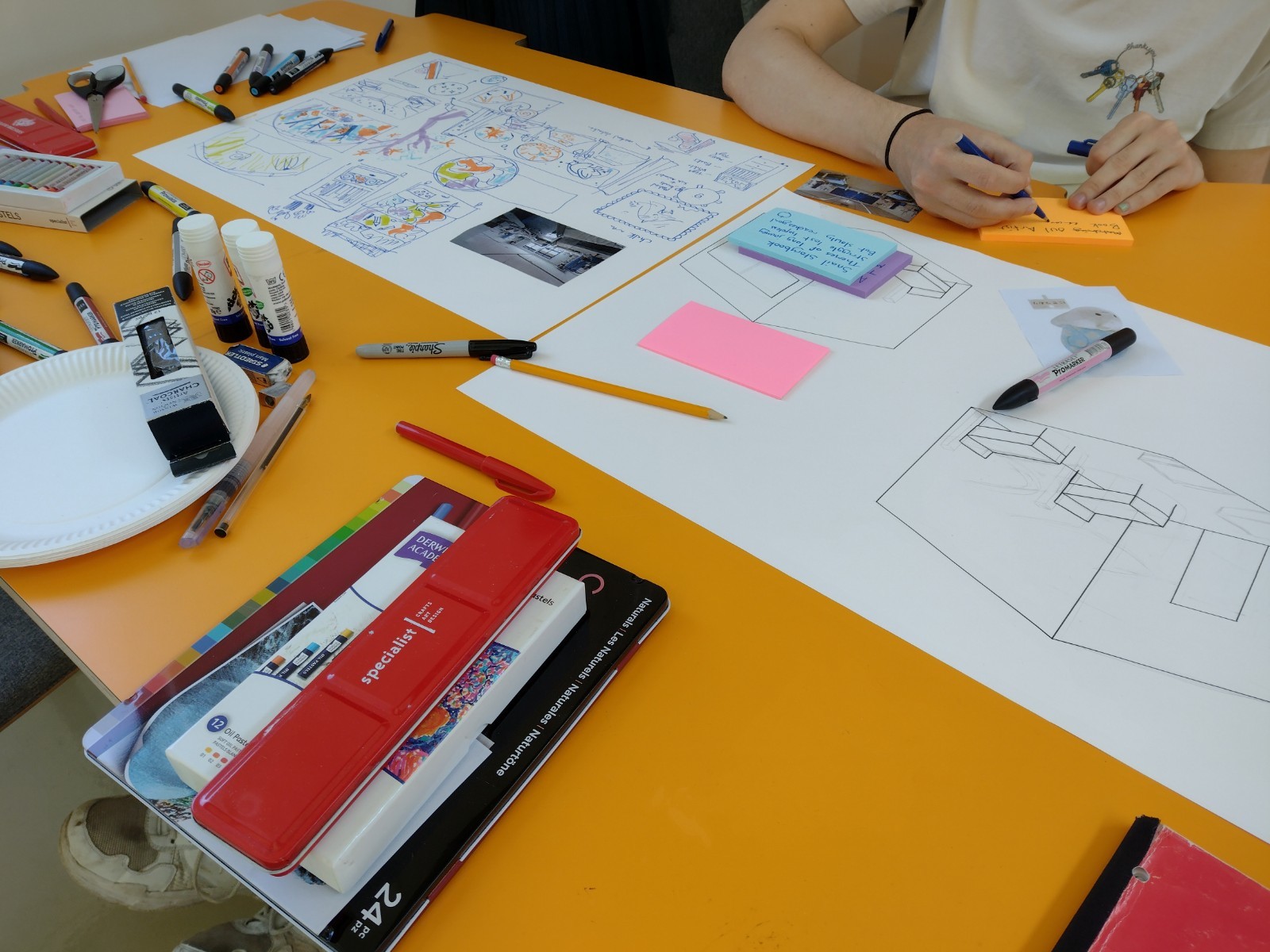 Two big sheets of paper with illustration projects on them. Someone sits at a table writing on a sticky note.