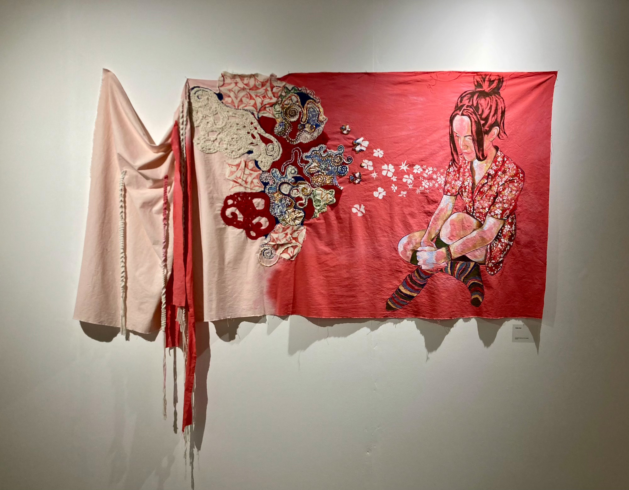 Large-scale, textile hanging displayed in the AUB gallery, depicting a loosely painted, seated young woman wearing a patterned floral dress and striped knee-high socks. The floral pattern flows off the dress onto the dyed red background, dispersing into intricate beaded embroidery, crochet, weaving and Lino cuts. Moving away from the figure, the dye fades and the corner of the piece is left draped and covered in macrame and fabric strands which hang below the bottom hem