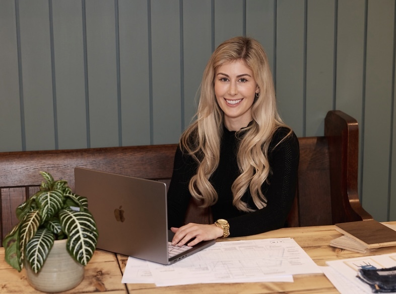 K Thomas sitting at wooden desk with laptop, papers, and potted plant against grey panelled wall.