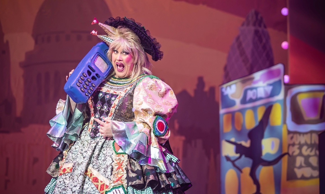 A performer dressed in a vibrant costume with a large blue mobile phone prop stands on stage. The background features colourful theatrical set pieces, contributing to the festive atmosphere of the performance.