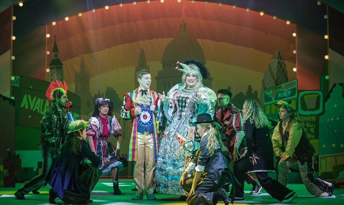 A group of performers on stage in a colourful theatrical production. The central figure, dressed in an elaborate costume, is surrounded by others in various outfits, with a vibrant cityscape backdrop and bright stage lighting.