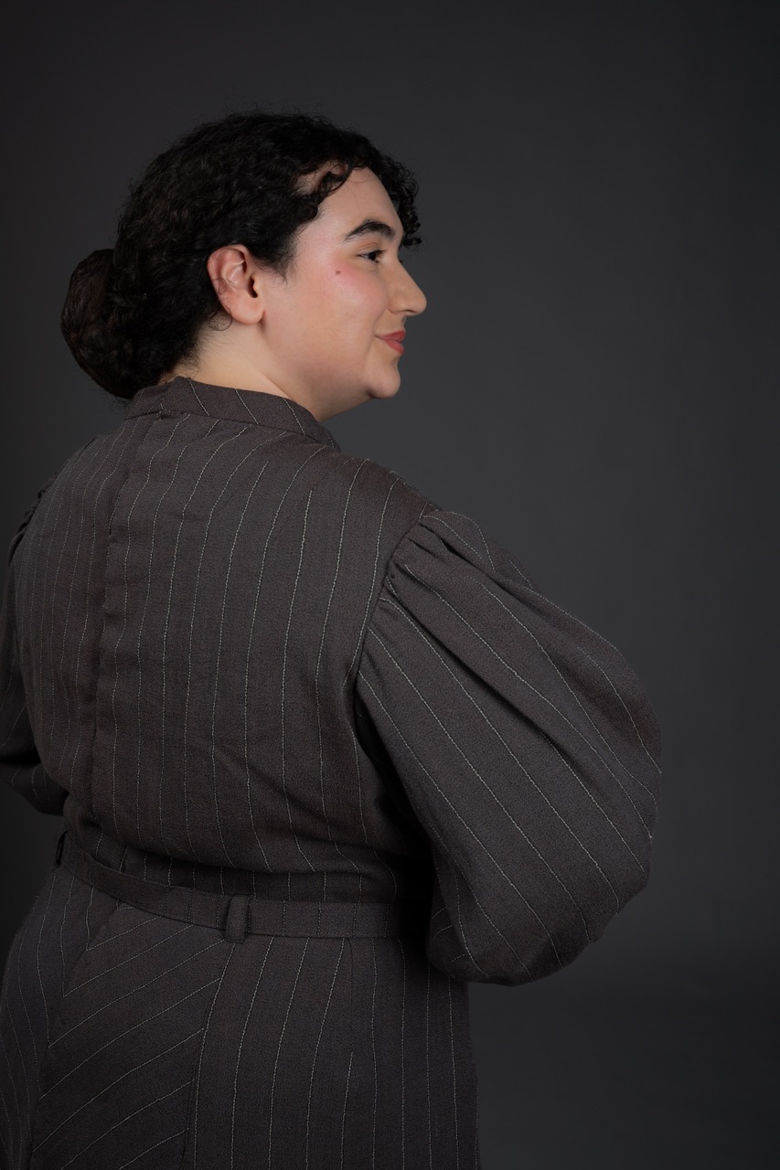 A woman with curly hair, dressed in a dark pinstriped outfit, stands with her back partially turned to the camera. The background is a solid dark colour, which contrasts with her clothing.