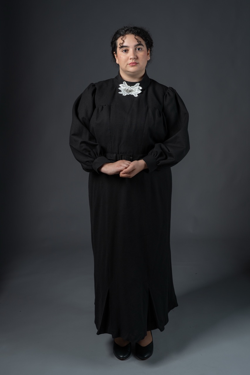 A woman stands upright in front of a grey backdrop, wearing a long black dress with puffed sleeves and a white collar. She has dark hair styled with curls and is looking directly at the camera.