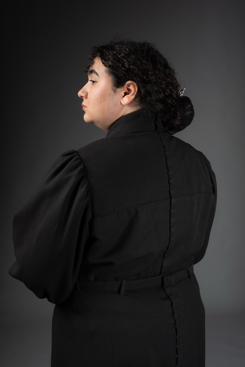 A person with curly dark hair is seen from the back, wearing a black high-collared garment with puffy sleeves. The photo is set against a dark grey background, highlighting the contours and details of the outfit.