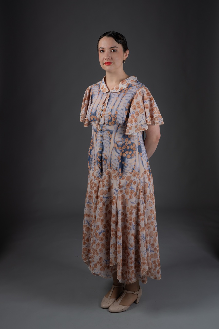 A woman with dark hair styled in a bun stands in profile, wearing a floral-patterned dress with short, ruffled sleeves. The dress features a light blue and beige colour scheme, and she stands against a dark background.