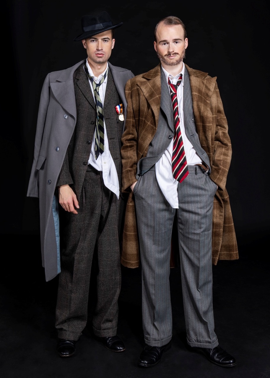 Two men in period costume: left wears dark suit with hat and medals, right in grey suit with brown overcoat and striped tie.