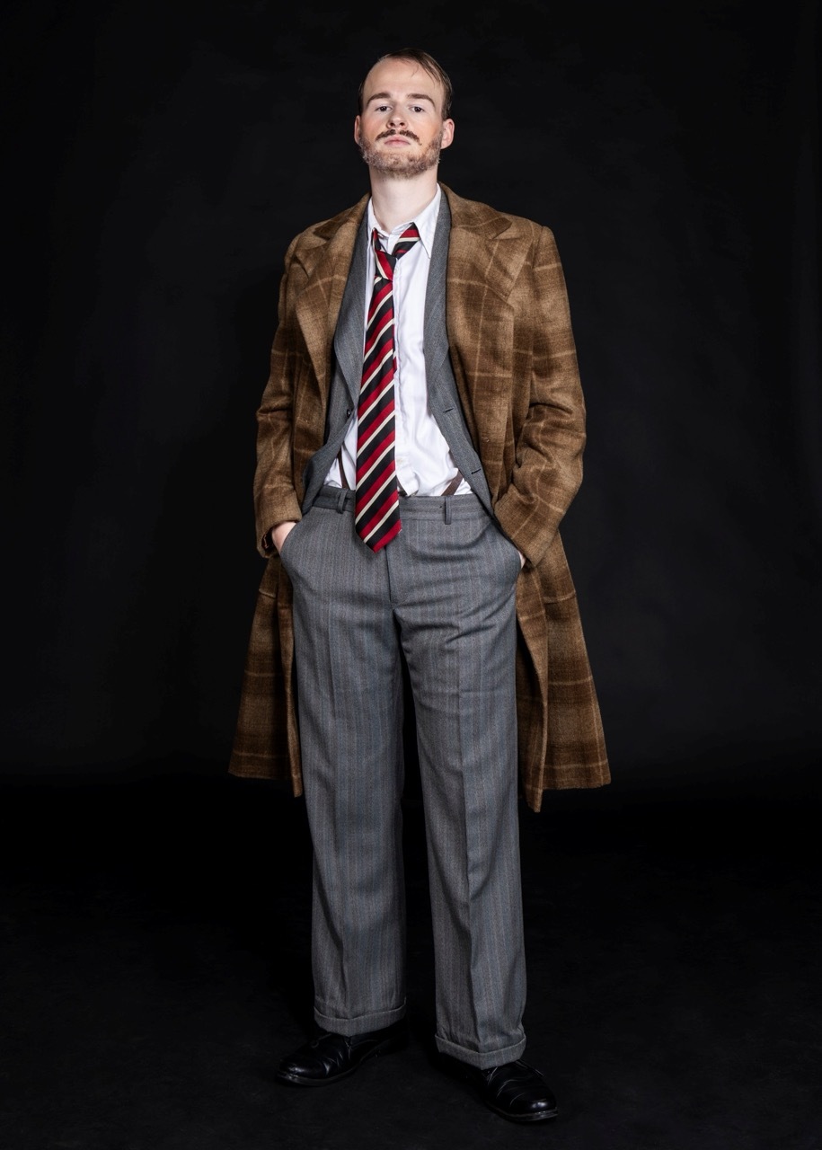 Man with moustache wearing brown overcoat, grey suit, white shirt and red striped tie, standing against black background.
