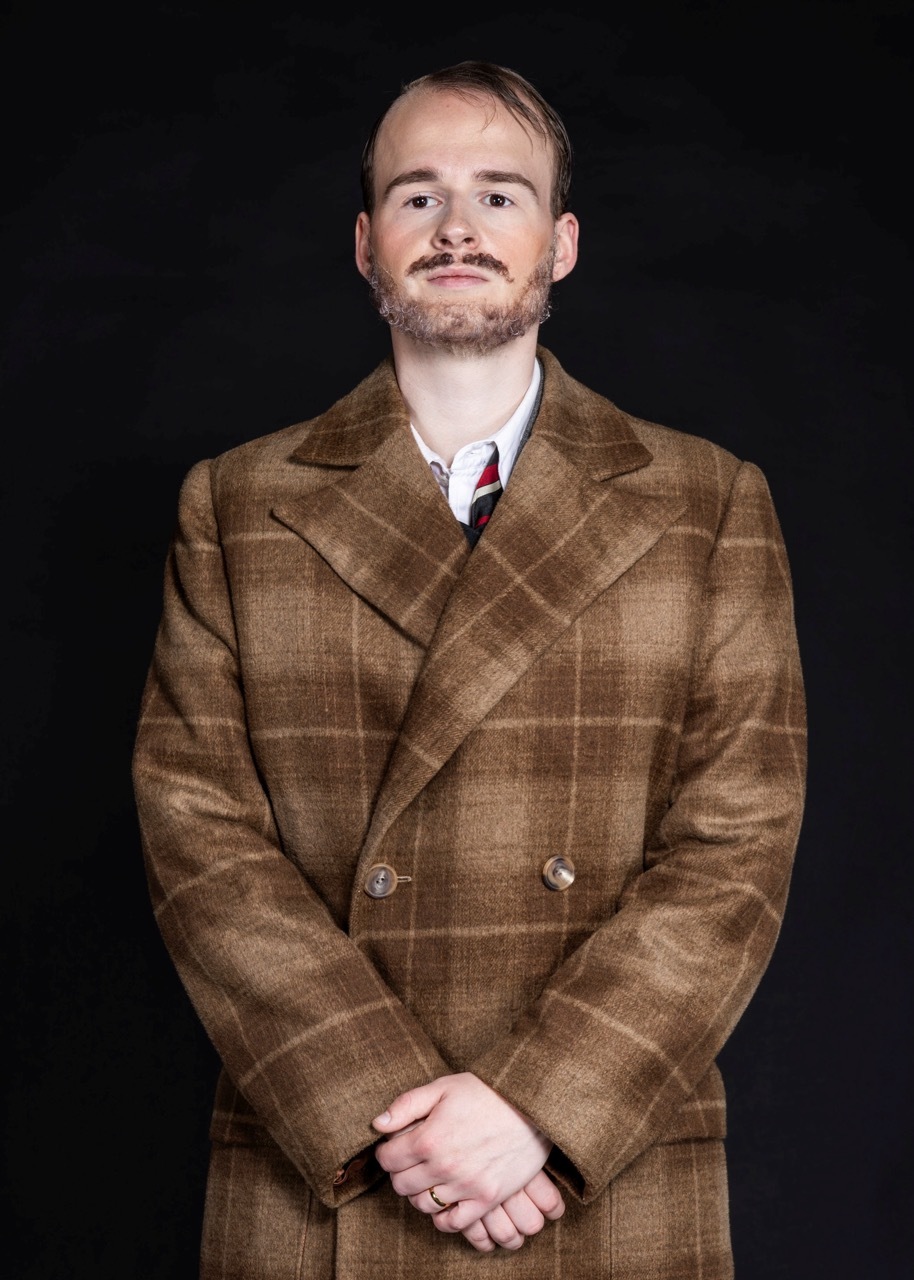 Man with beard wearing brown tweed double-breasted coat, white shirt and red tie against black background