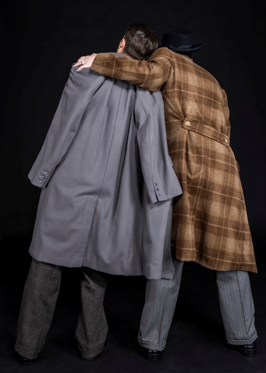 Two men embracing, one in grey suit, other in brown checked coat, against dark background.