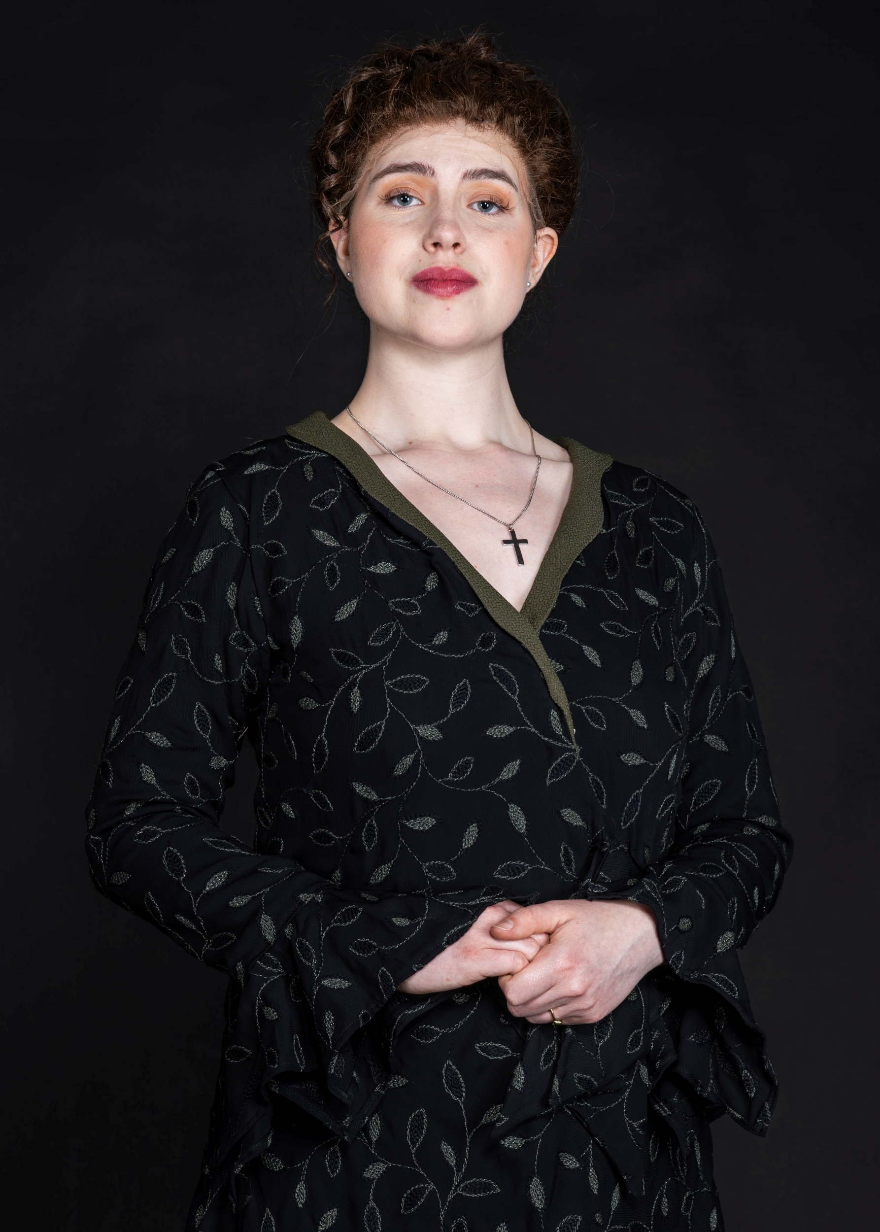 Woman with curly brown hair wearing dark floral dress with green trim and cross necklace, hands clasped, against black background.