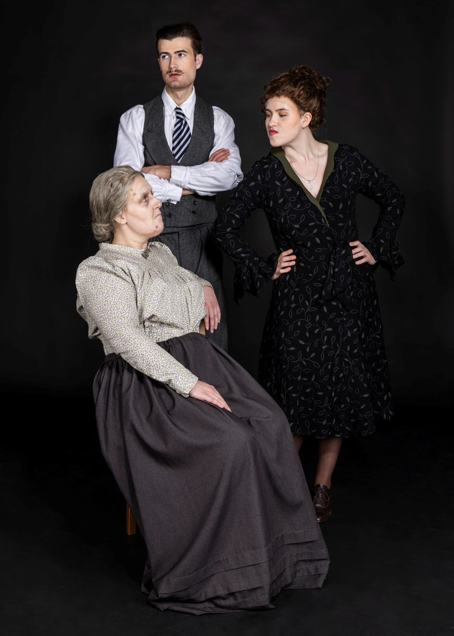 Three actors in period costume against black backdrop: seated woman in grey dress, standing man in waistcoat with crossed arms, woman in black dress with hands on hips.