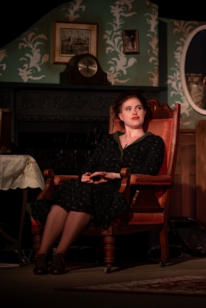 Woman in black dress sitting in wooden chair beside fireplace with green floral wallpaper, framed pictures, and mantel clock.
