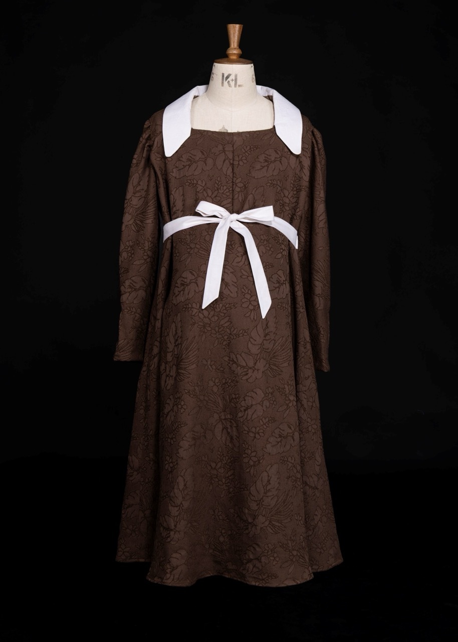 Brown textured dress with white collar and sleeves, white ribbon sash at waist, displayed on mannequin against black background.